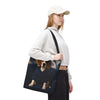 A woman wearing a white sweatshirt, black pants, and a white cap carries a dark tote bag featuring a printed image of a beagle dog in a blue hoodie, set against a plain white background.