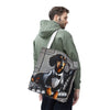 A man wearing an olive green hoodie is shown from the back, carrying a large tote bag over his shoulder. The tote bag features a printed graphic of a dachshund dressed in a black tuxedo and bow tie, sitting on a gray tufted leather couch.