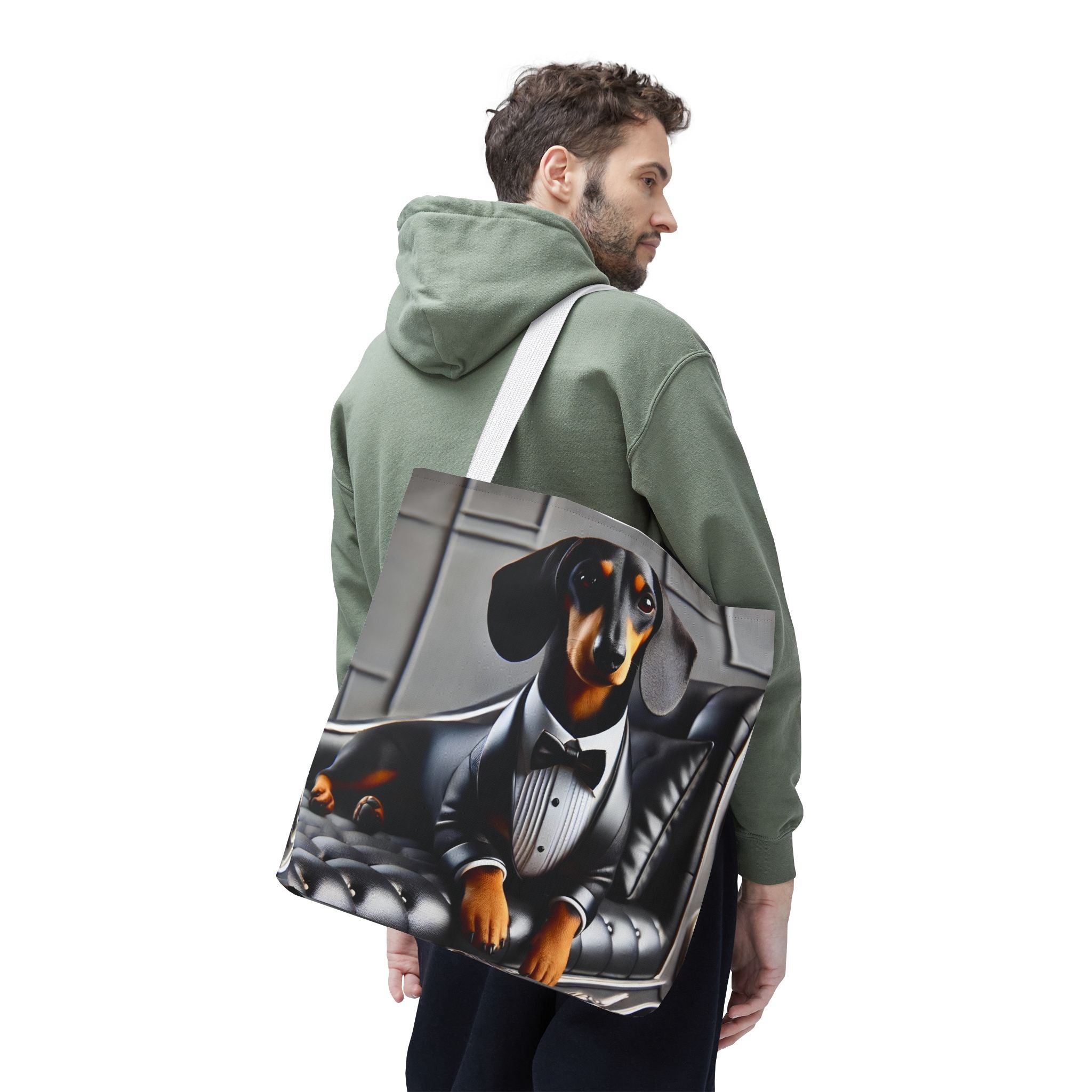A man wearing an olive green hoodie is shown from the back, carrying a large tote bag over his shoulder. The tote bag features a printed graphic of a dachshund dressed in a black tuxedo and bow tie, sitting on a gray tufted leather couch.