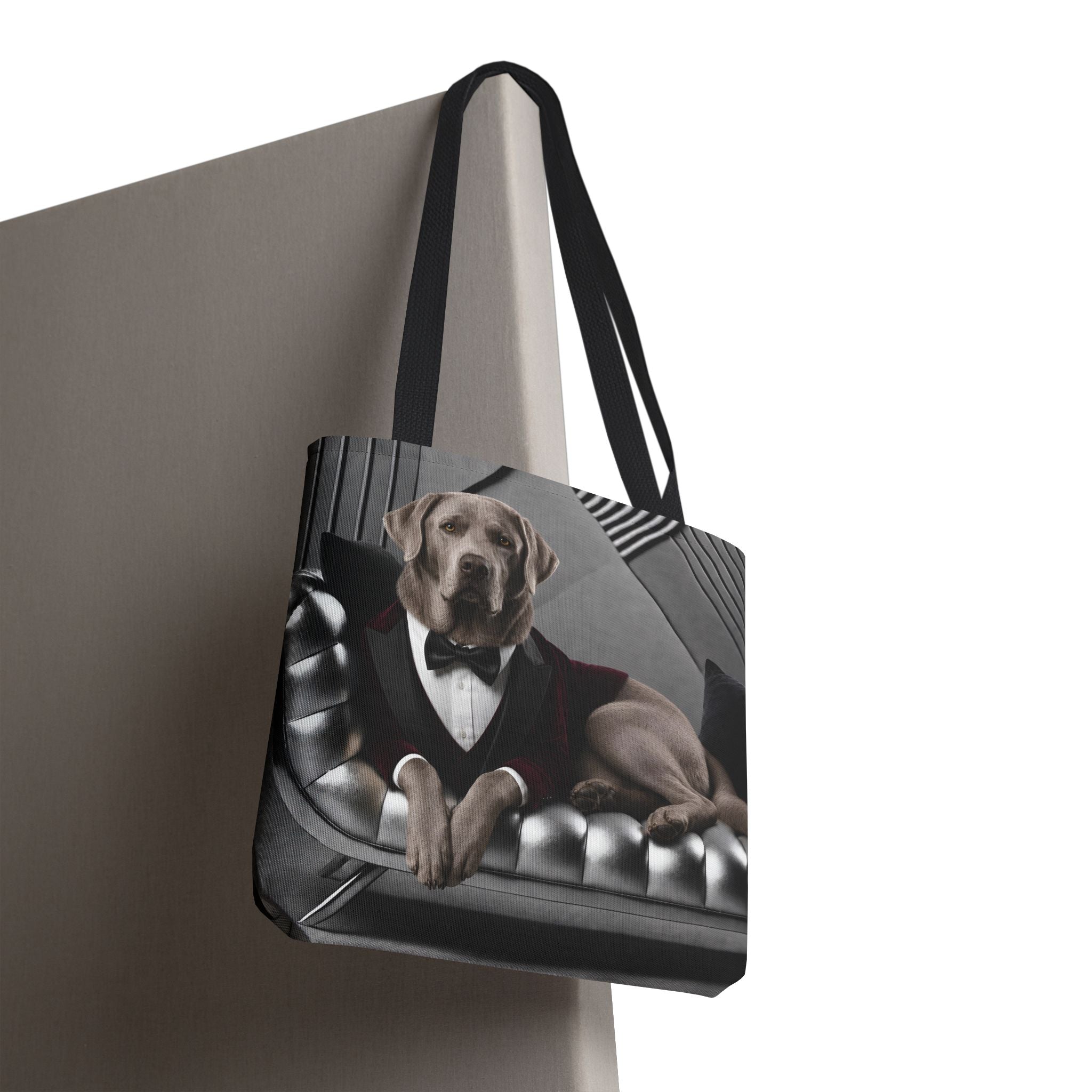 A tote bag with black handles, featuring a printed graphic of a silver Labrador retriever dressed in a tuxedo and bowler hat, sitting on a tufted grey sofa. The bag is shown hanging against a neutral grey wall on a white background.