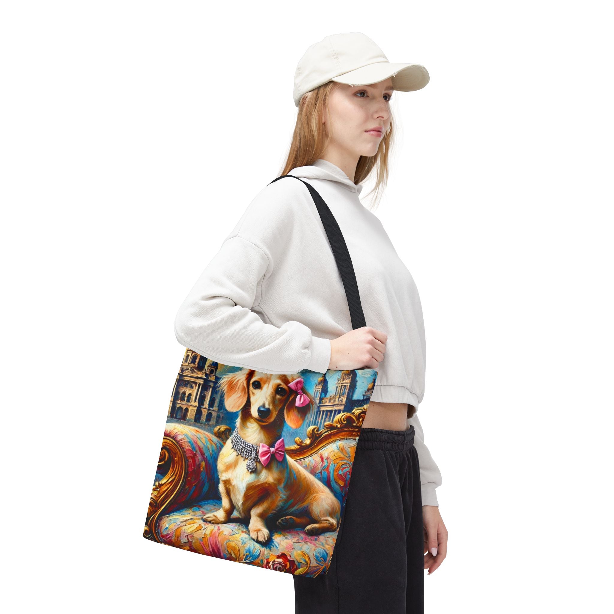 A woman wearing a white cap, white sweatshirt, and black pants carries a canvas tote bag featuring a colorful painting of a dachshund on a baroque chair.