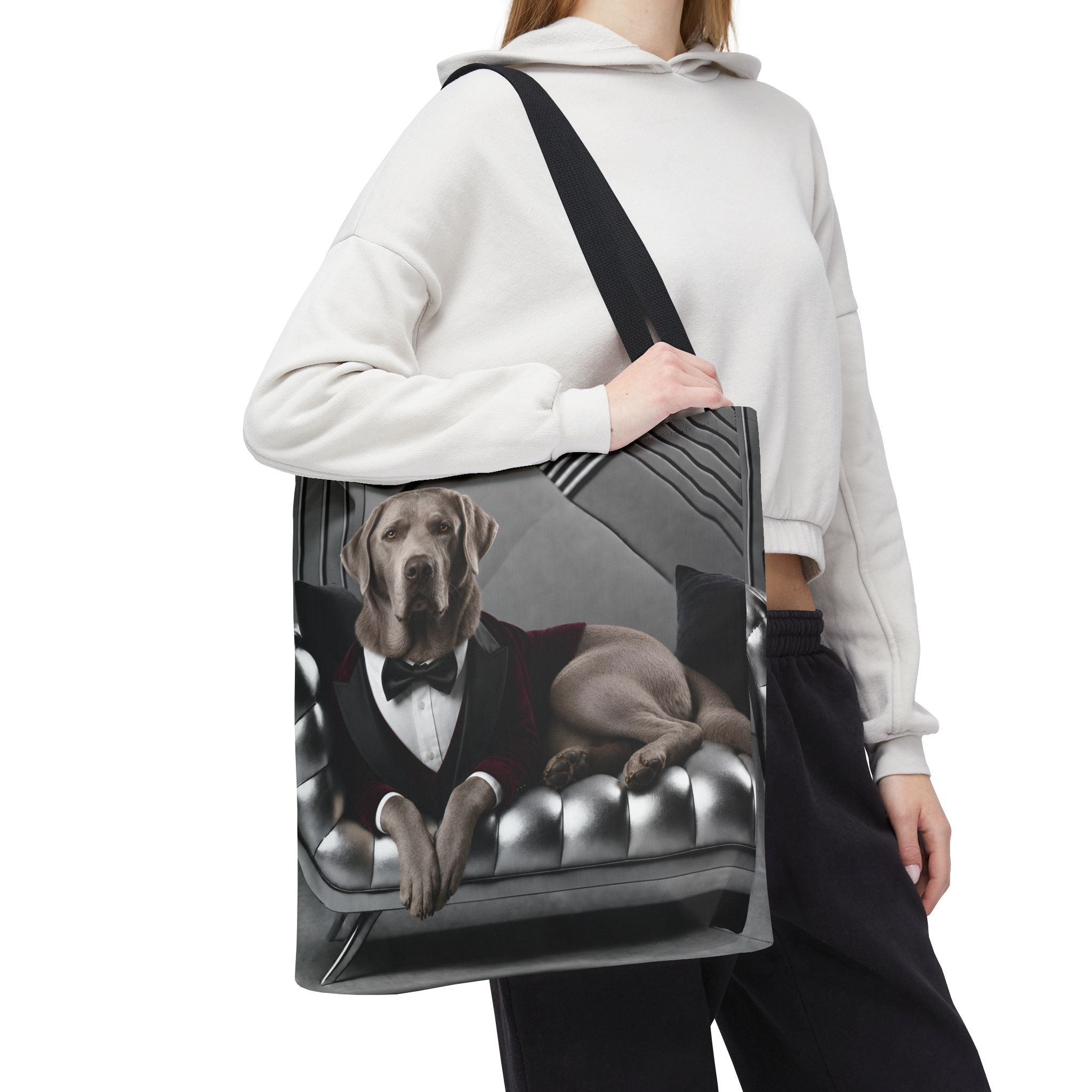 A person carries a large tote bag over their shoulder. The tote bag features a printed graphic of a silver Labrador retriever dressed in a black tuxedo sitting on a tufted gray sofa.