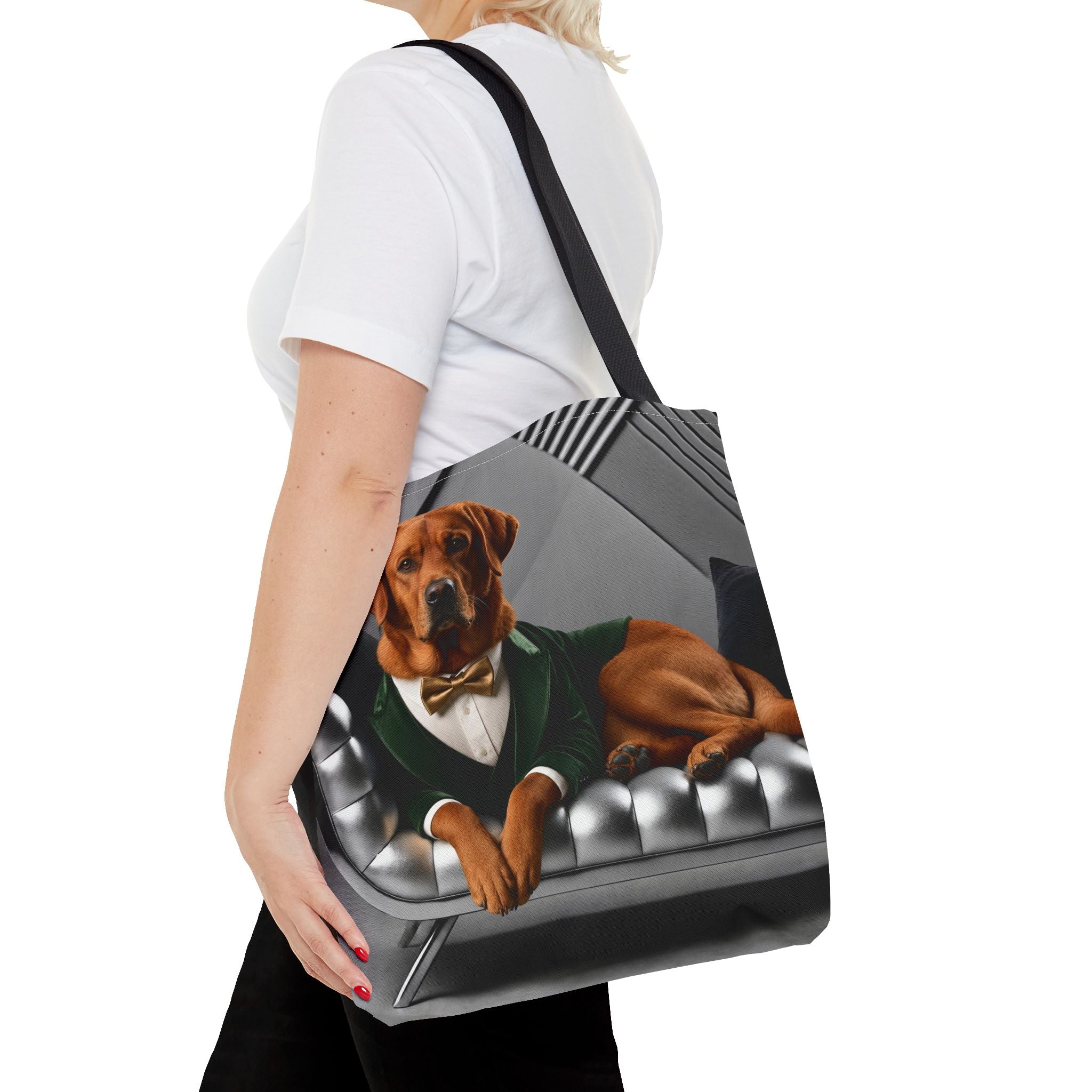 A woman carries a tote bag over her shoulder. The tote bag features a printed graphic of a fox red Labrador retriever dressed in a green suit and bow tie, lying on a tufted gray sofa.