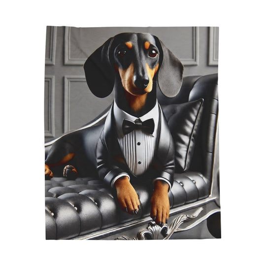 A blanket featuring a dachshund dressed in a black tuxedo with a white shirt and bow tie, sitting on a black tufted chaise lounge against a gray paneled wall.