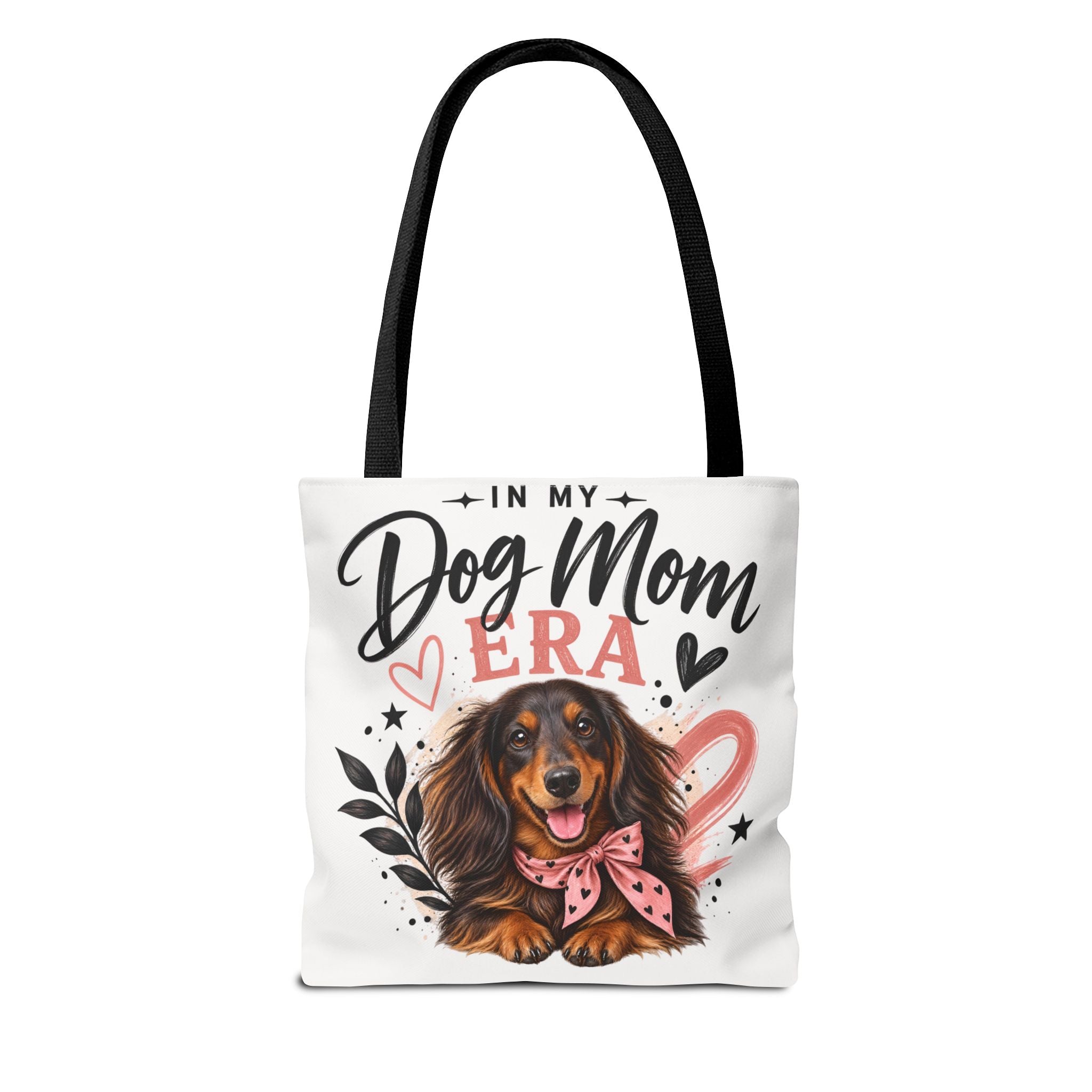 A white canvas tote bag with black shoulder straps, featuring a printed graphic of a dachshund wearing a pink bow, surrounded by the text 'IN MY Dog Mom ERA' with hearts and stars.