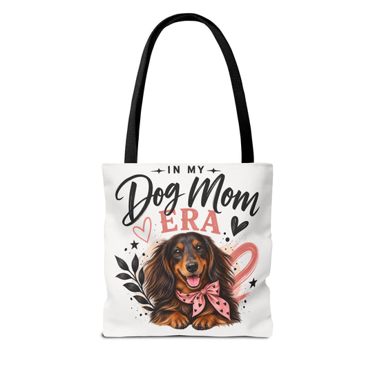 A white canvas tote bag with black shoulder straps, featuring a printed graphic of a dachshund wearing a pink bow, surrounded by the text 'IN MY Dog Mom ERA' with hearts and stars.