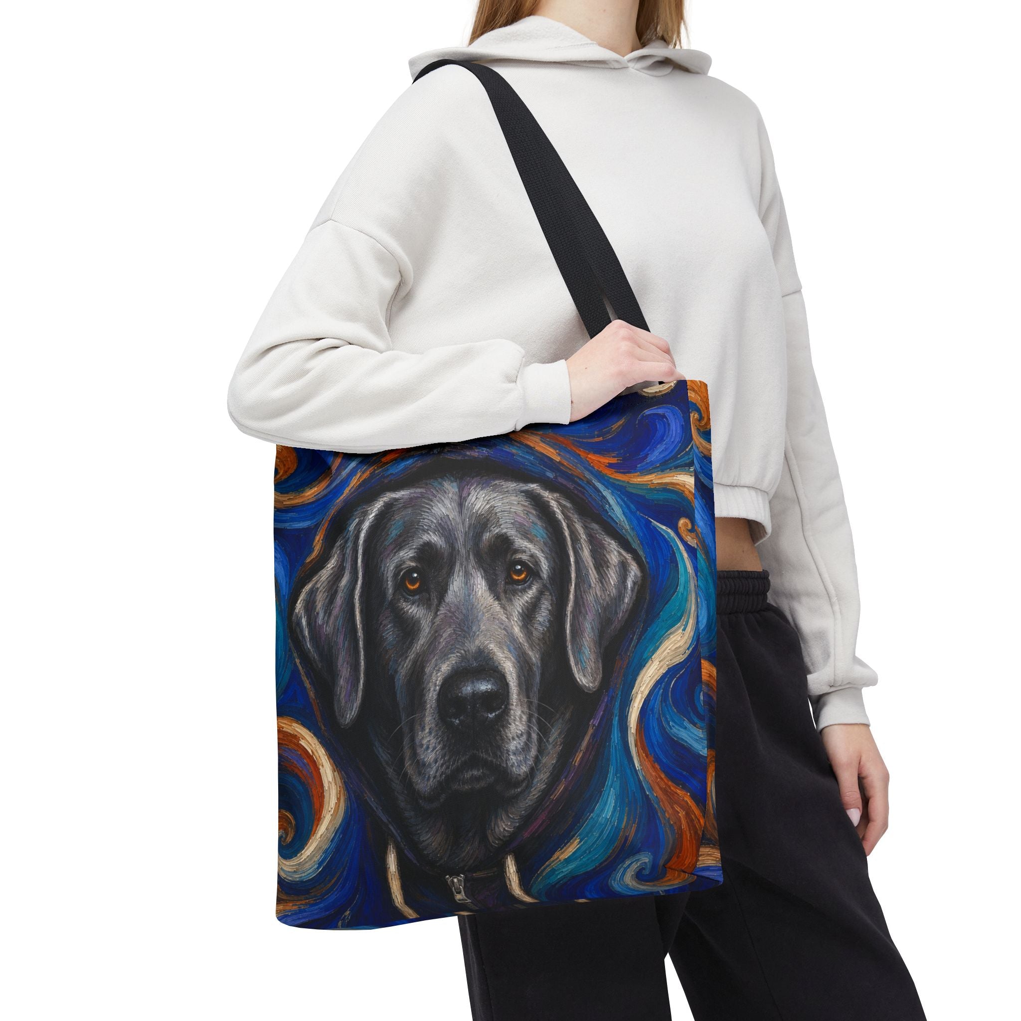 A person wearing a white cropped hoodie and black sweatpants carries a canvas tote bag featuring a painted silver Labrador retriever portrait against a swirling blue, orange, and black background.