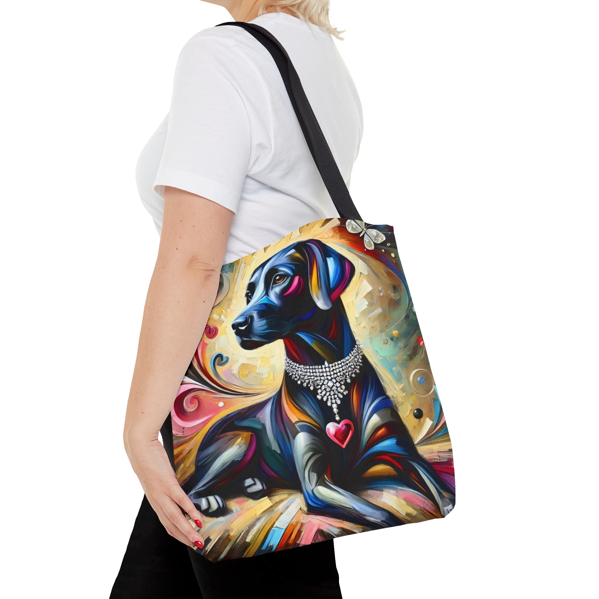 A woman wearing a white t-shirt and black pants carries a colorful tote bag featuring a stylized painting of a black Labrador retriever with a silver collar and a pink heart, set against a plain white background.