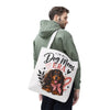 A man wearing a sage green hoodie and black pants is shown from the back, carrying a white canvas tote bag over his shoulder. The bag features a printed illustration of a long-haired dachshund wearing a pink bow tie, with the text 'In My Dog Mom ERA' and small red hearts.
