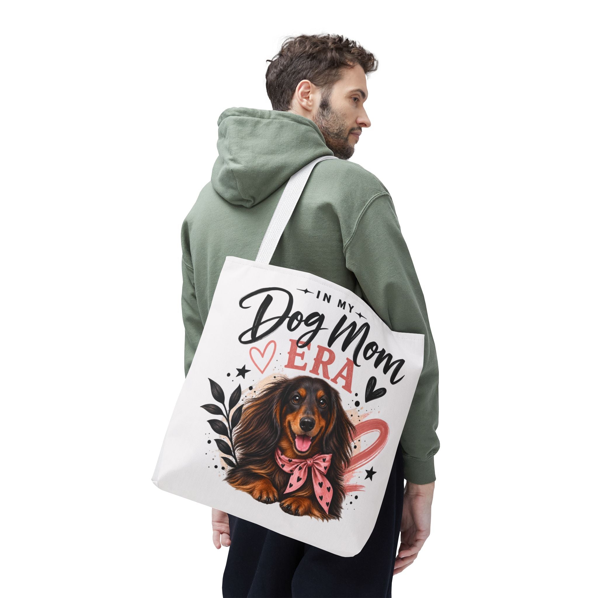 A man wearing a sage green hoodie and black pants is shown from the back, carrying a white canvas tote bag over his shoulder. The bag features a printed illustration of a long-haired dachshund wearing a pink bow tie, with the text 'In My Dog Mom ERA' and small red hearts.