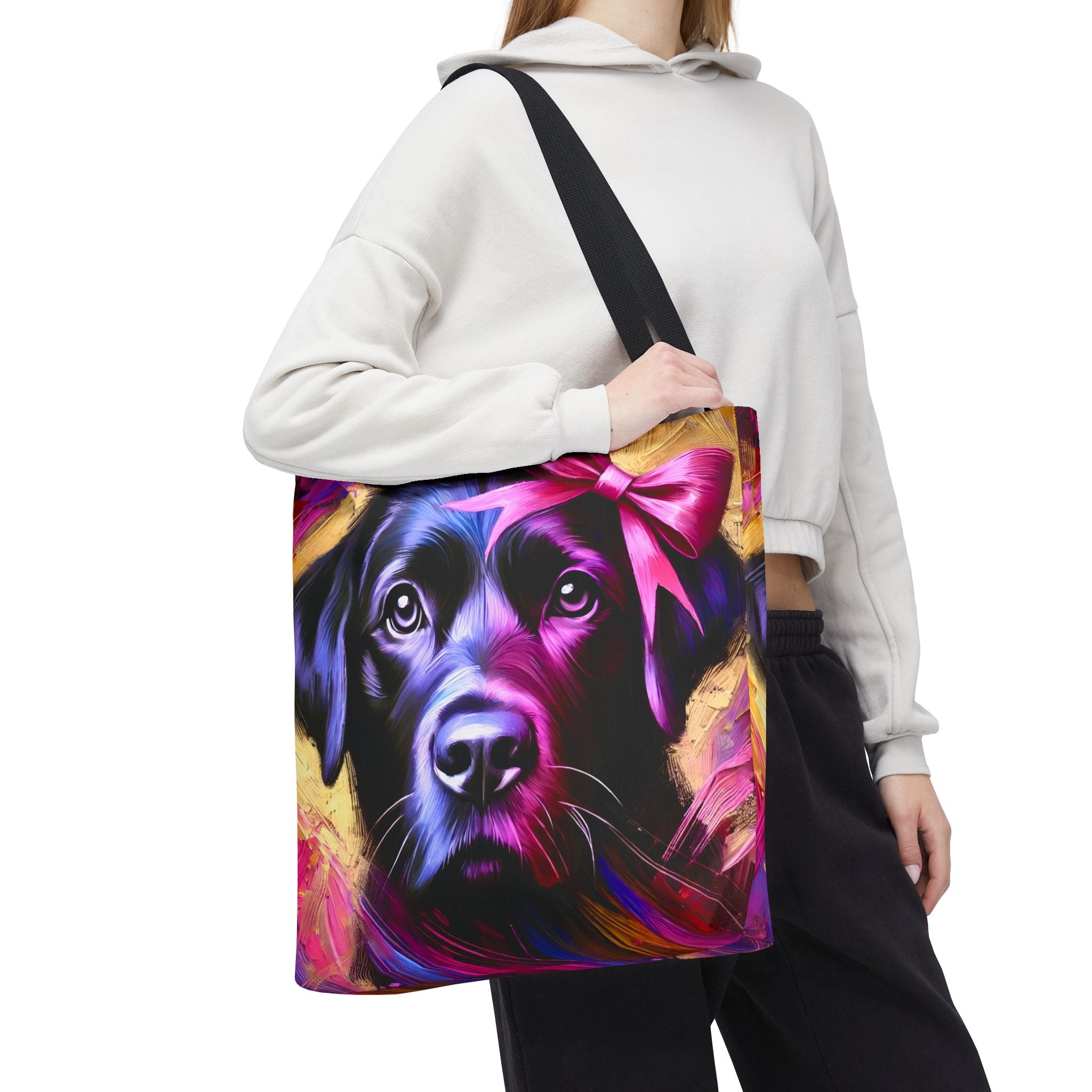 A person wearing a white cropped hoodie and black sweatpants carries a canvas tote bag with a colorful graphic print of a black Labrador wearing a pink bow, against a plain white background.