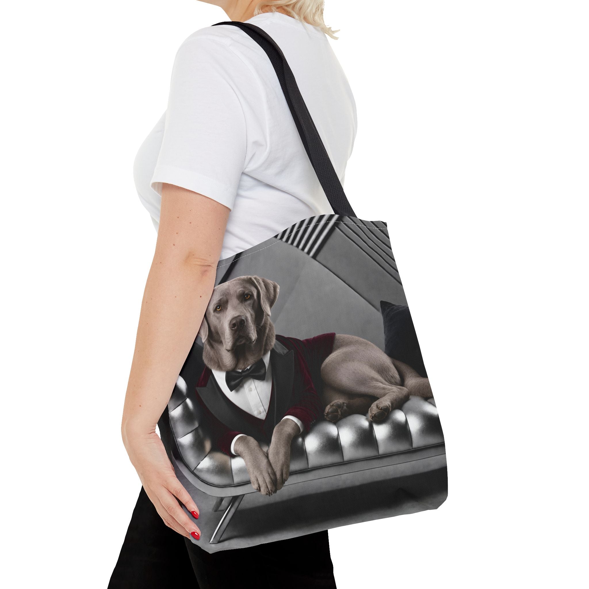 A woman carries a black shoulder tote bag featuring a printed graphic of a silver Labrador retriever dressed in a tuxedo sitting in a tufted grey sofa.