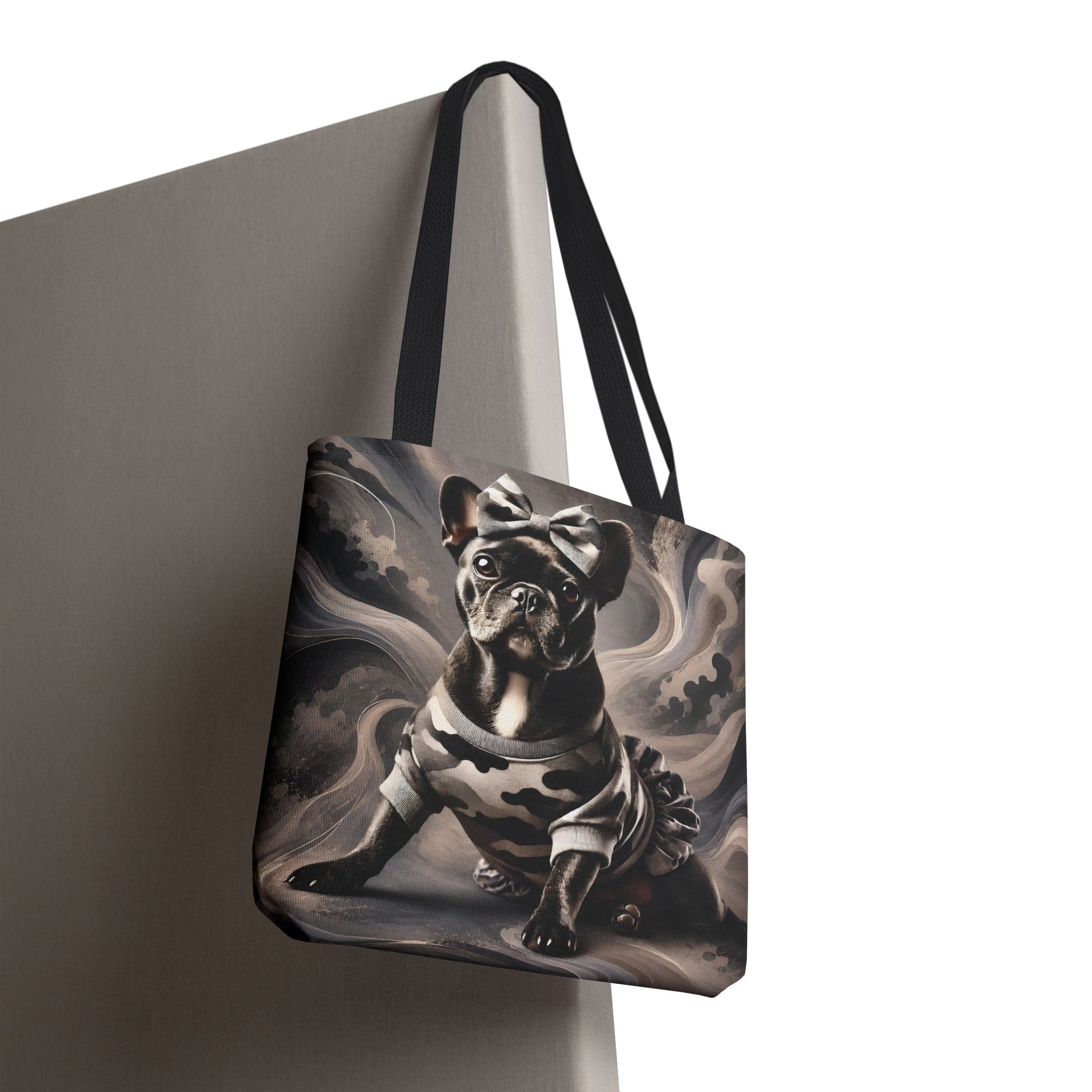 A tote bag with black shoulder straps, featuring a printed graphic of a black French Bulldog wearing a bow and a ruffled dress, set against a swirling gray and black background. The bag is shown hanging from a gray support structure against a white background.