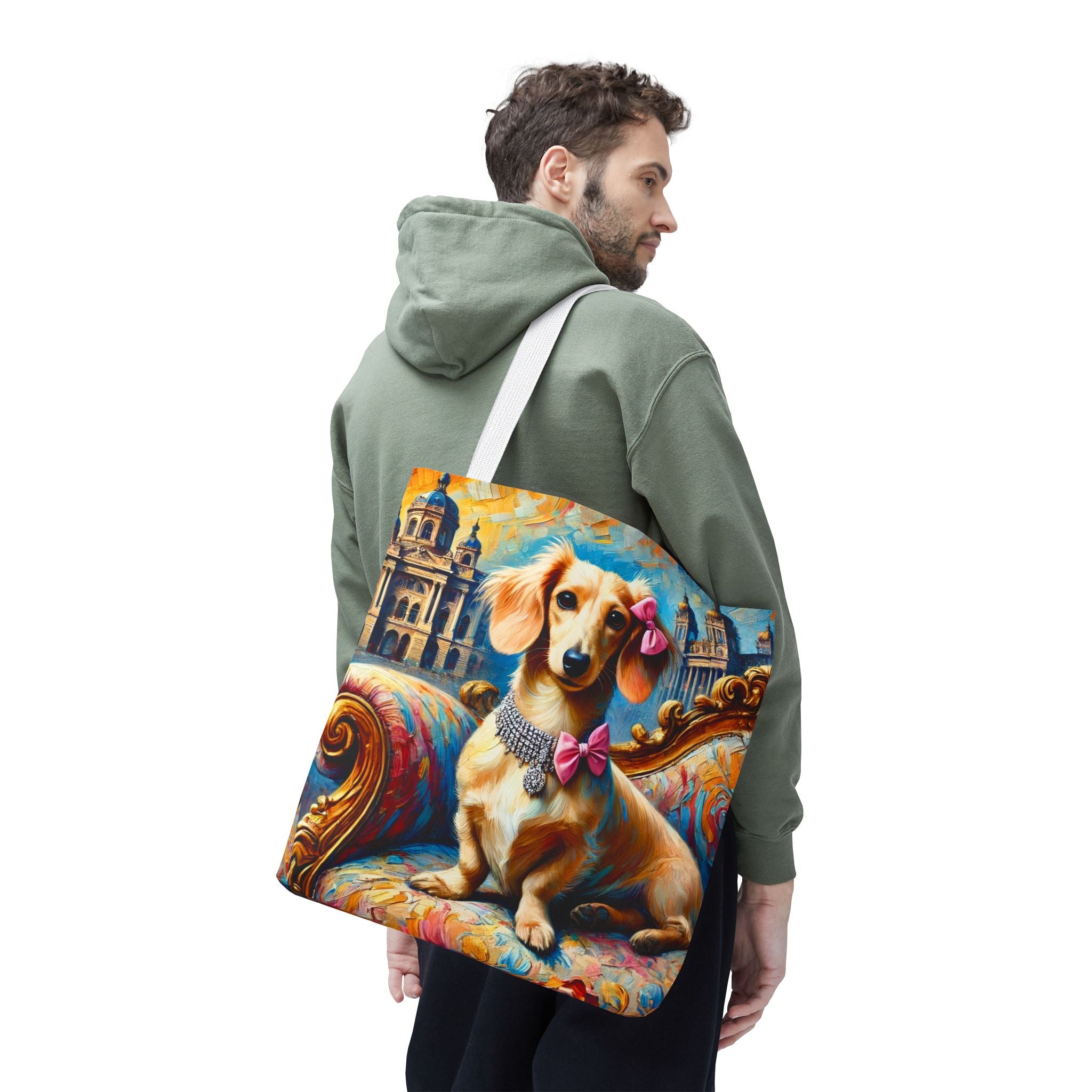 A man wearing an olive green hoodie carries a canvas tote bag over his shoulder. The bag features a colorful, artistic print of a dachshund wearing a pink bow on an ornate chaise lounge with a blue and gold background.
