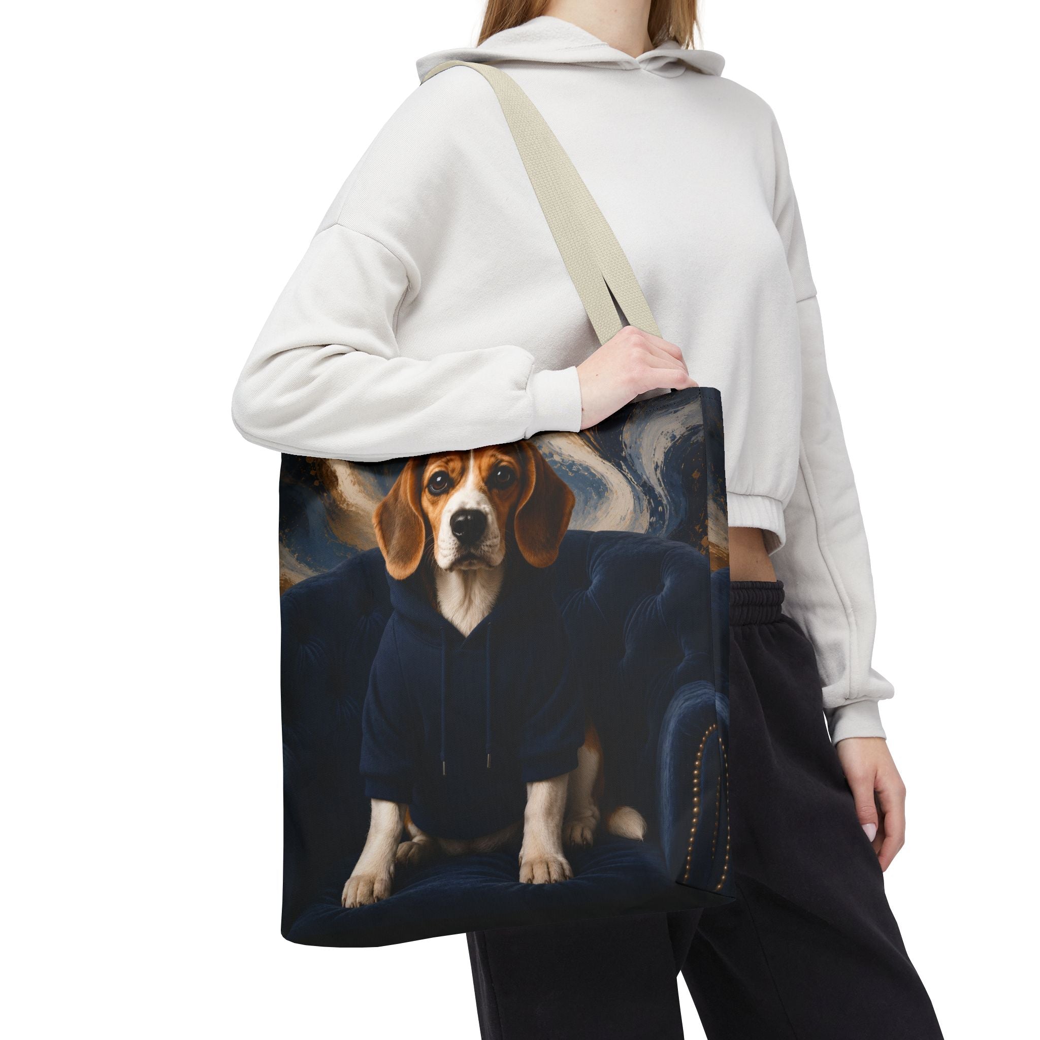 A person wearing a white cropped hoodie and black sweatpants carries a canvas tote bag over their shoulder. The tote bag features a printed graphic of a beagle dog sitting on a dark blue armchair against a swirling dark background.