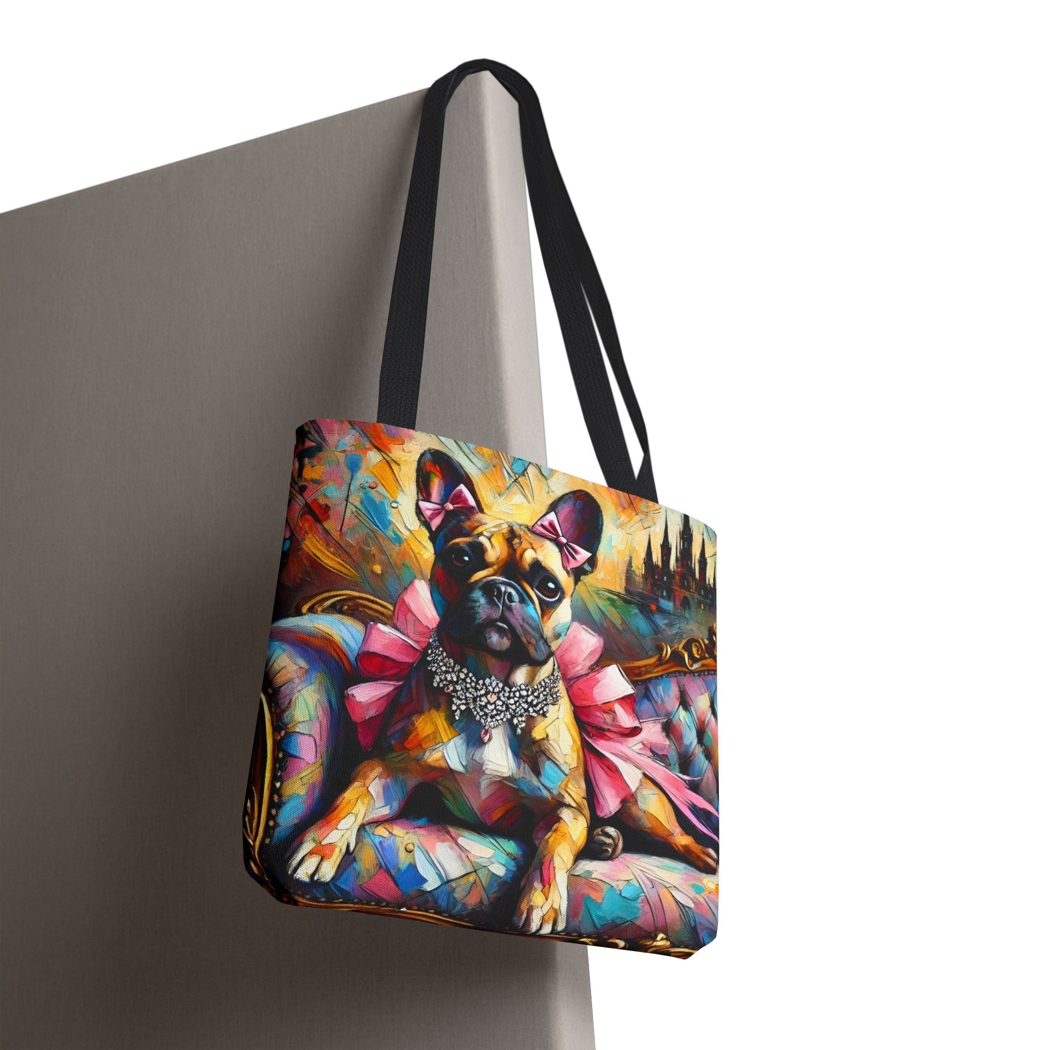 A tote bag with black shoulder straps, featuring a colorful, painterly illustration of a French Bulldog wearing pink bows and a silver collar, shown hanging against a gray wall on a white background.