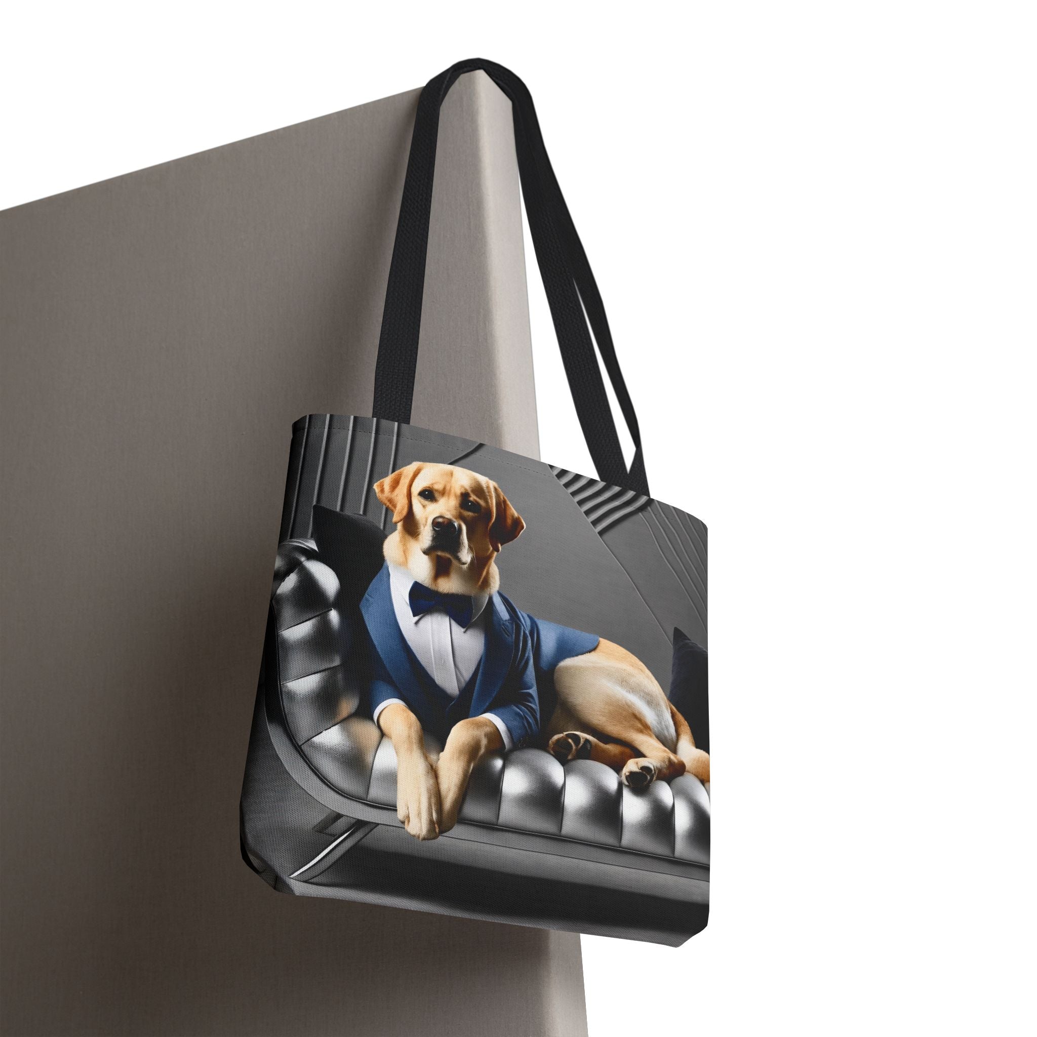 A tote bag with black handles, featuring a printed graphic of a yellow Labrador retriever wearing a blue suit and bow tie while lying on a black tufted leather sofa. The bag is shown hanging against a neutral gray background.