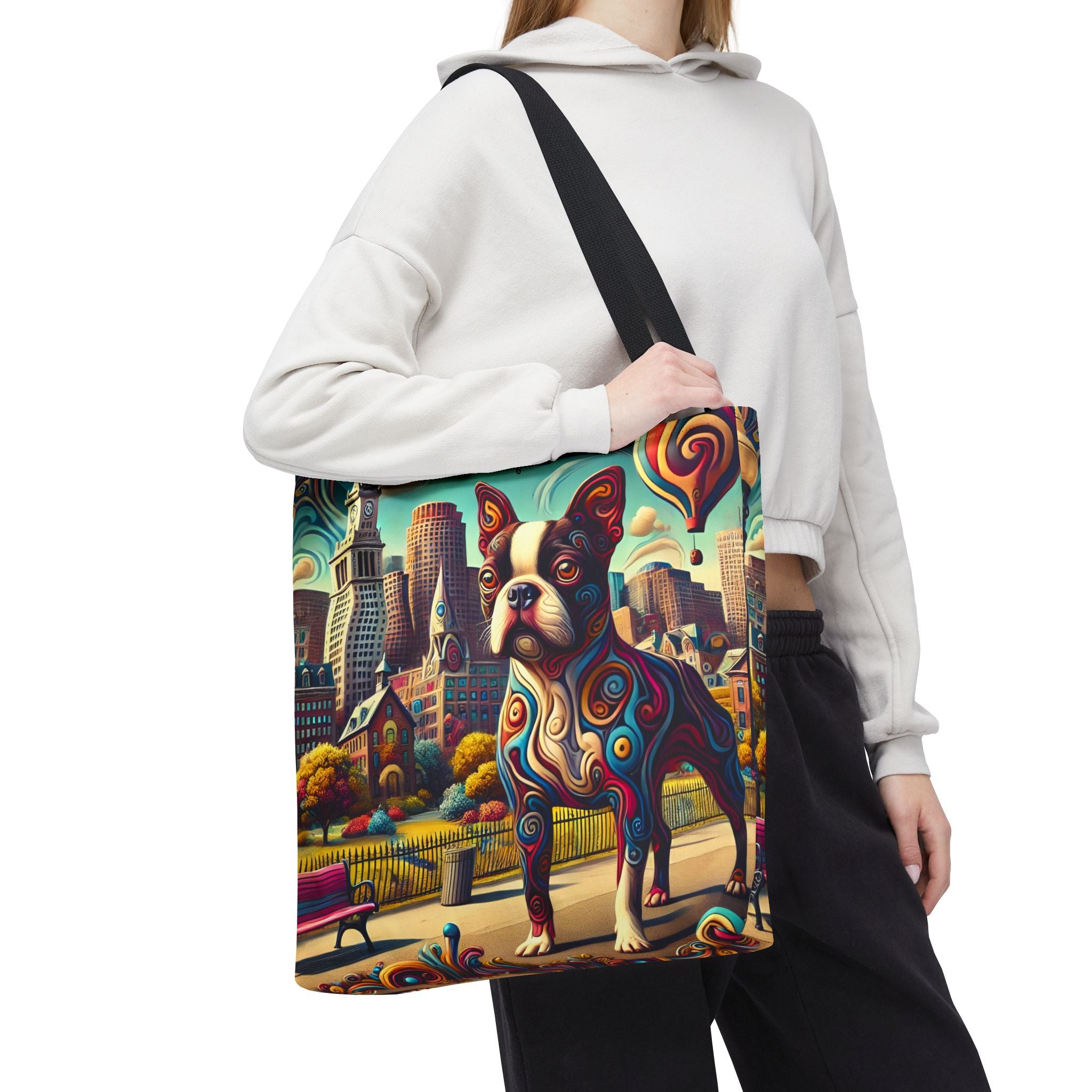 A person wearing a white hoodie and black sweatpants carries a large tote bag over their shoulder. The tote bag features a colorful, artistic print of a Boston Terrier dog in front of a city skyline, with a black shoulder strap.