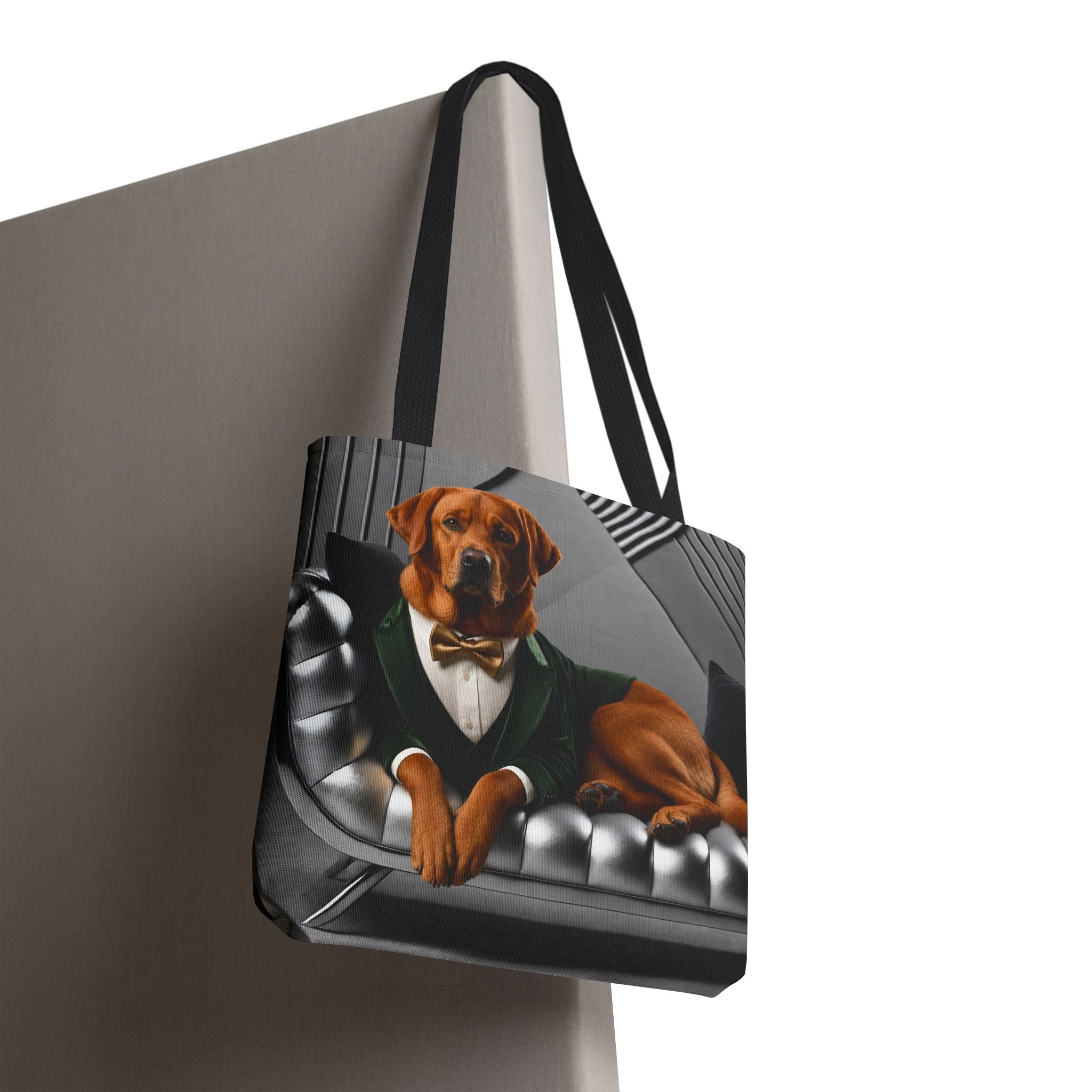 A tote bag with black handles, featuring a printed graphic of a fox red Labrador retriever dressed in a black tuxedo with a white shirt and bow tie, sitting on a black tufted sofa. The bag is shown hanging against a neutral gray background.