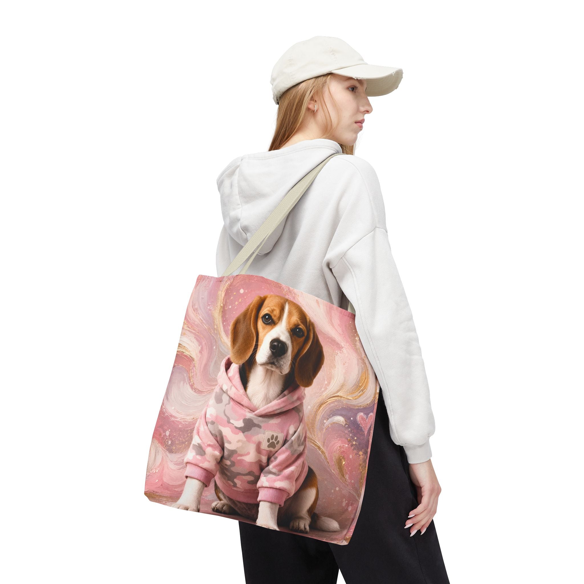 A woman wearing a white cap and white hoodie carries a canvas tote bag with a printed illustration of a beagle dog in a pink hoodie against a pink and gold abstract background.