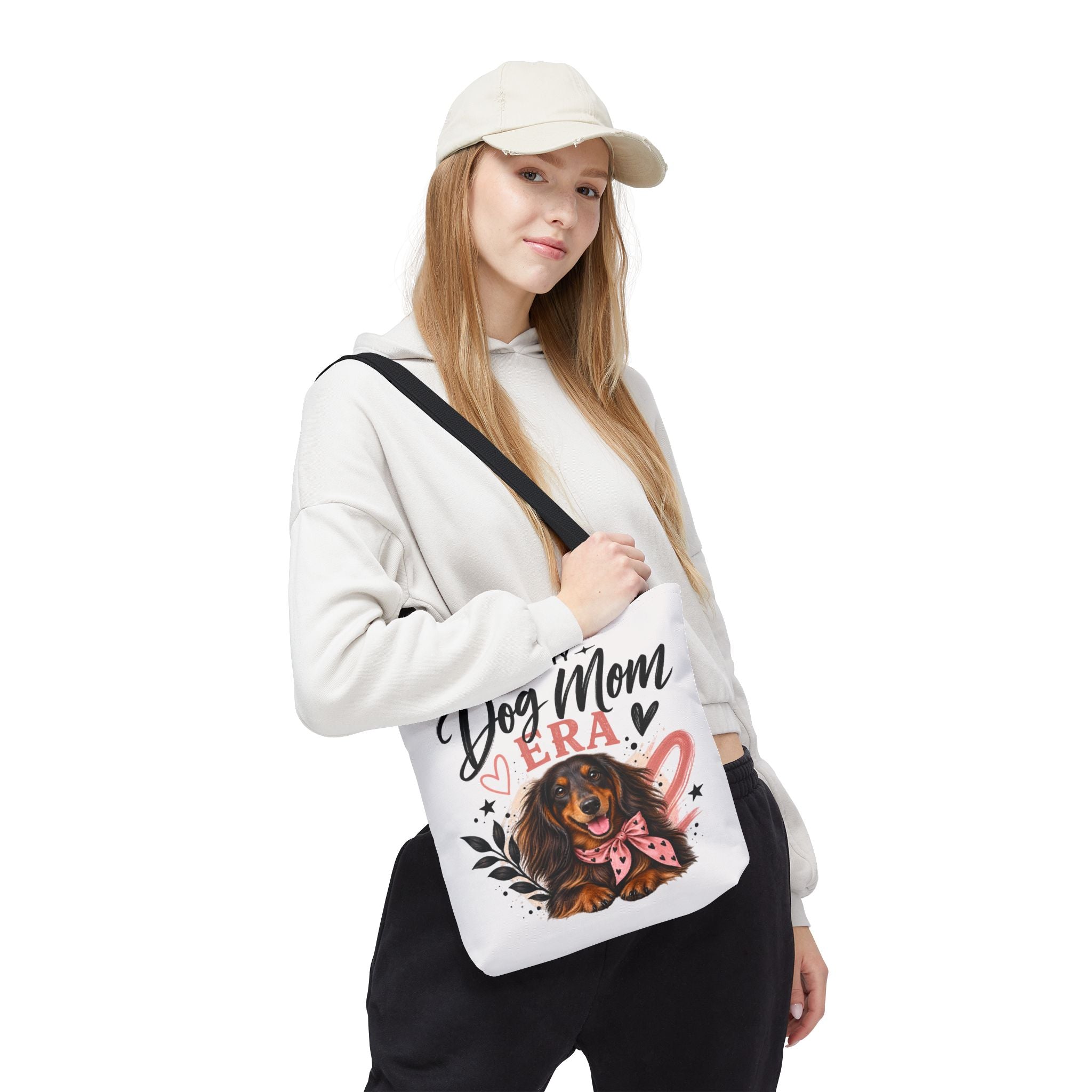 A woman wearing a beige cap, white hoodie, and black sweatpants carries a white canvas shoulder bag featuring a dachshund illustration and the text 'Dog Mom ERA.
