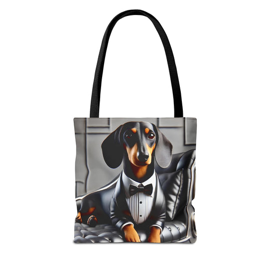 A tote bag with black shoulder straps, featuring a printed graphic of a dachshund dressed in a black tuxedo and bow tie, sitting on a gray tufted chair against a light gray background.