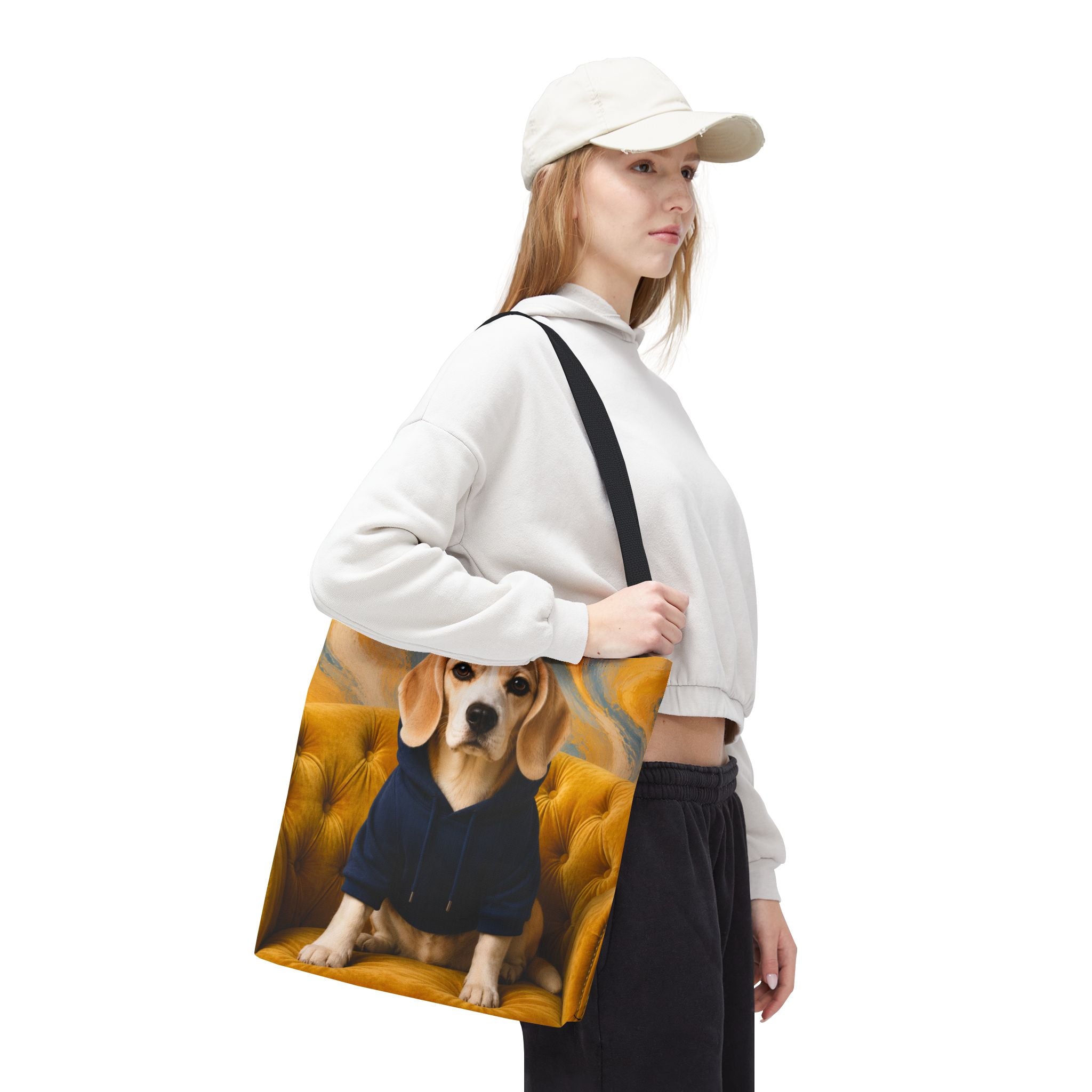 A woman wearing a white cap, white sweatshirt, and black pants carries a canvas tote bag featuring a printed portrait of a beagle dog sitting on a tufted yellow chair.