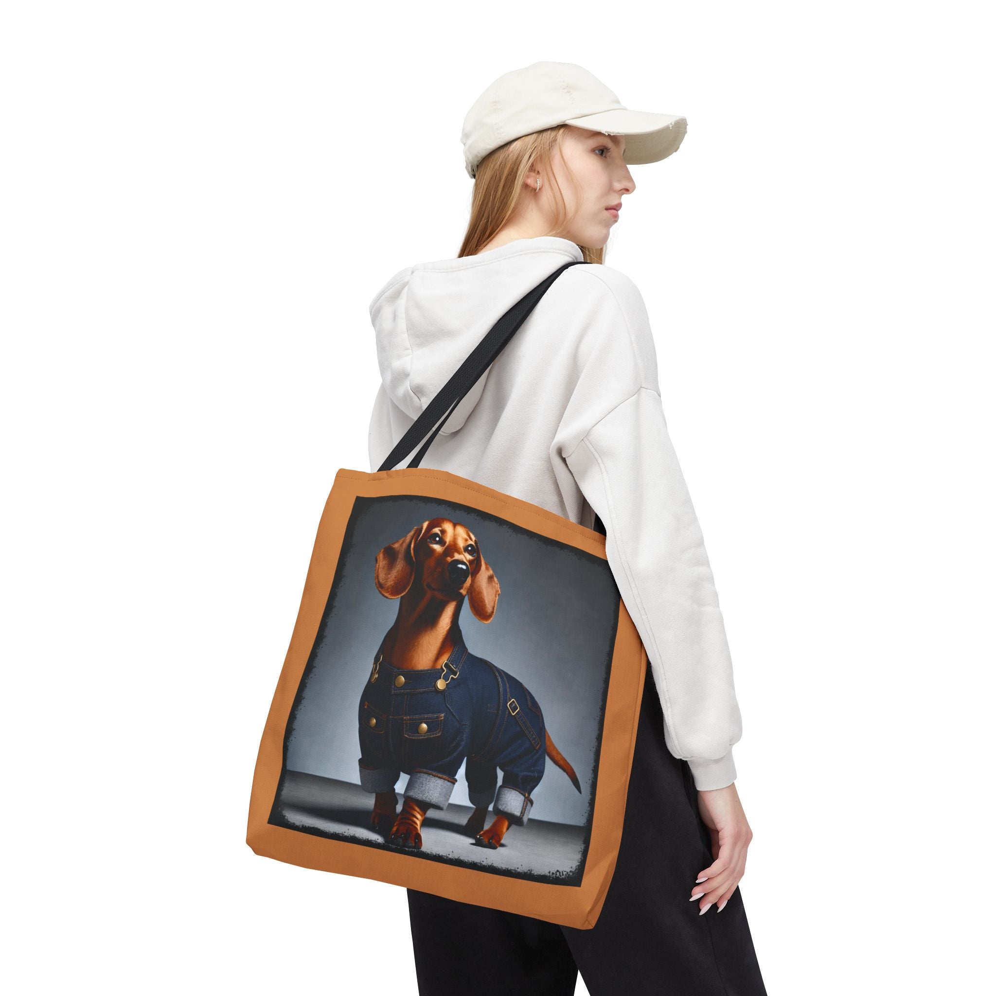 A woman wearing a white cap and white hoodie carries a canvas tote bag with a printed dachshund wearing denim overalls. The bag has a brown leather-look border and a black shoulder strap, shown against a plain white background.