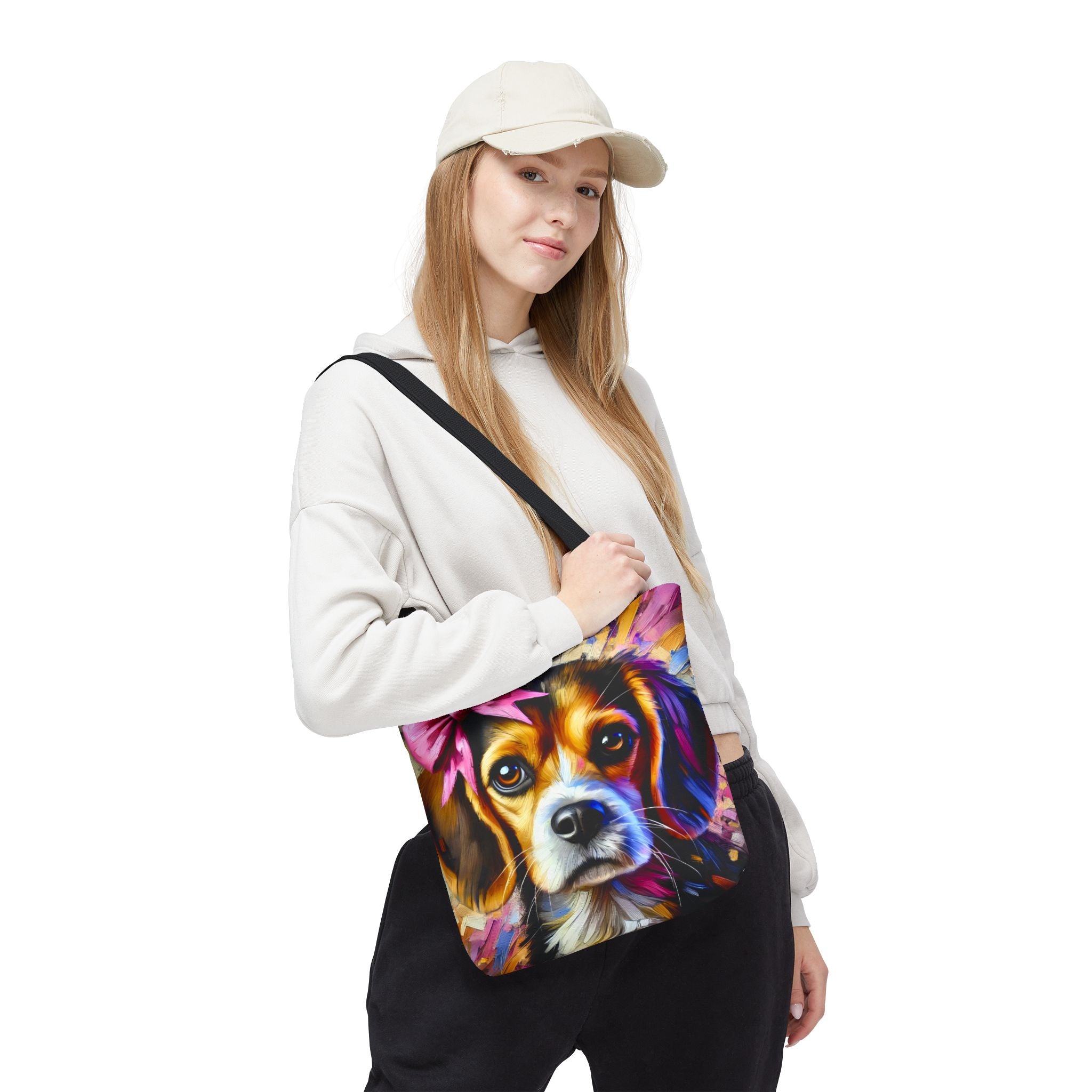 A woman wearing a beige cap, white hoodie, and black sweatpants carries a canvas tote bag with a colorful, vibrant painted beagle portrait on the front. The background is solid white.