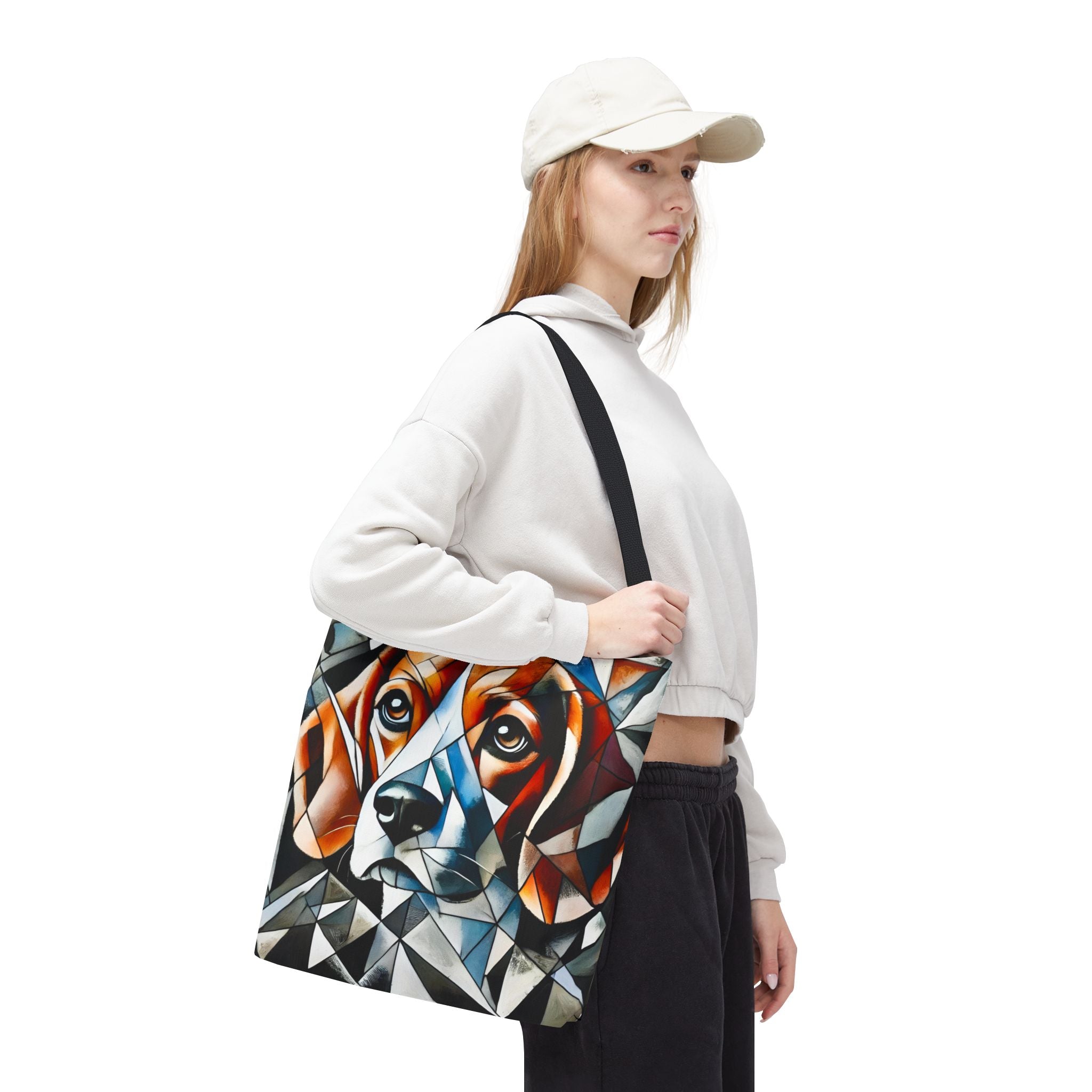 A woman wearing a white hoodie, black sweatpants, and a cream baseball cap is shown from the side, carrying a large canvas tote bag. The bag features a modern geometric art print of a beagle's  face in shades of orange, brown, black, white, and blue.