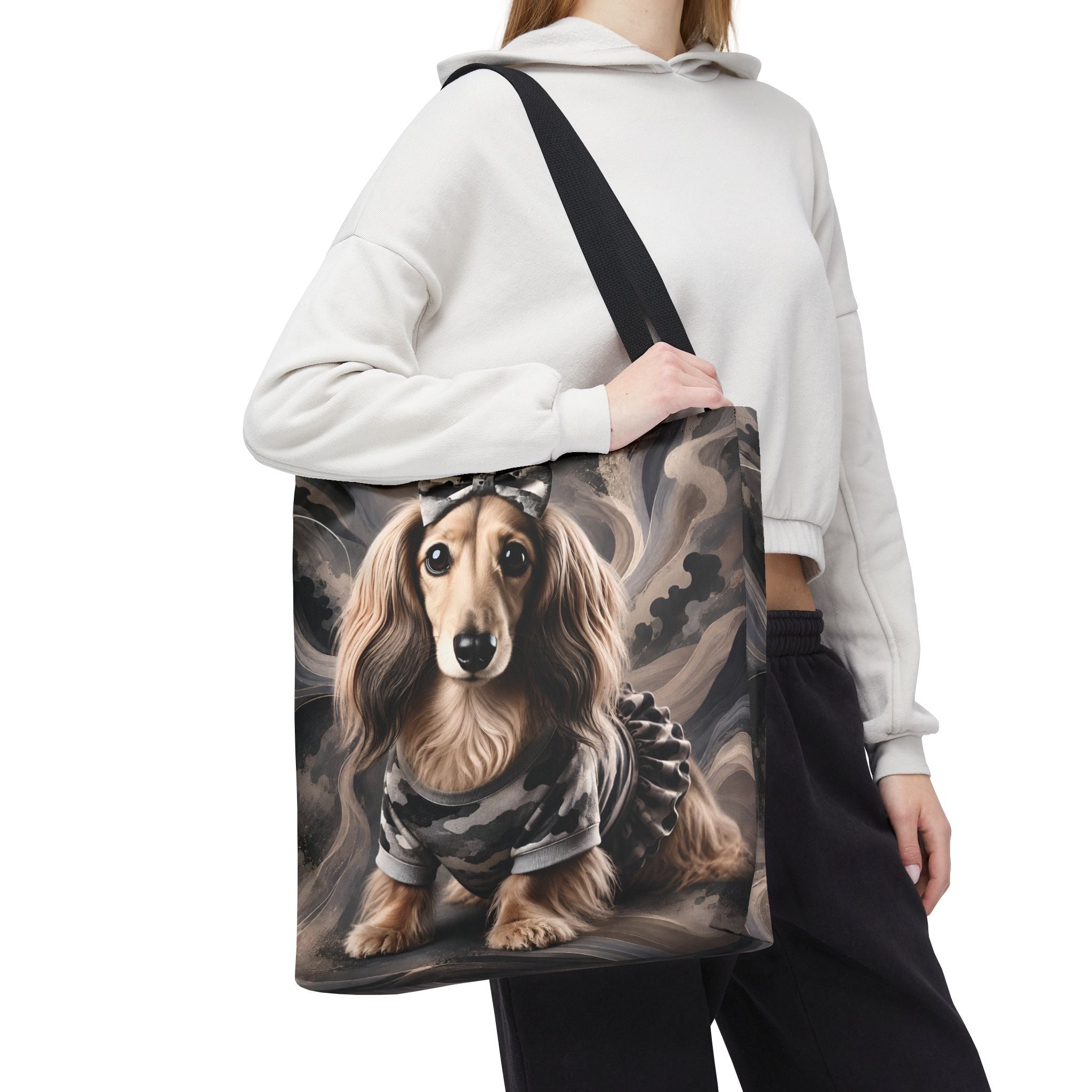A person wearing a white cropped hoodie and black sweatpants carries a large tote bag featuring a printed dachshund dog in a camouflage outfit against a smoky gray background.
