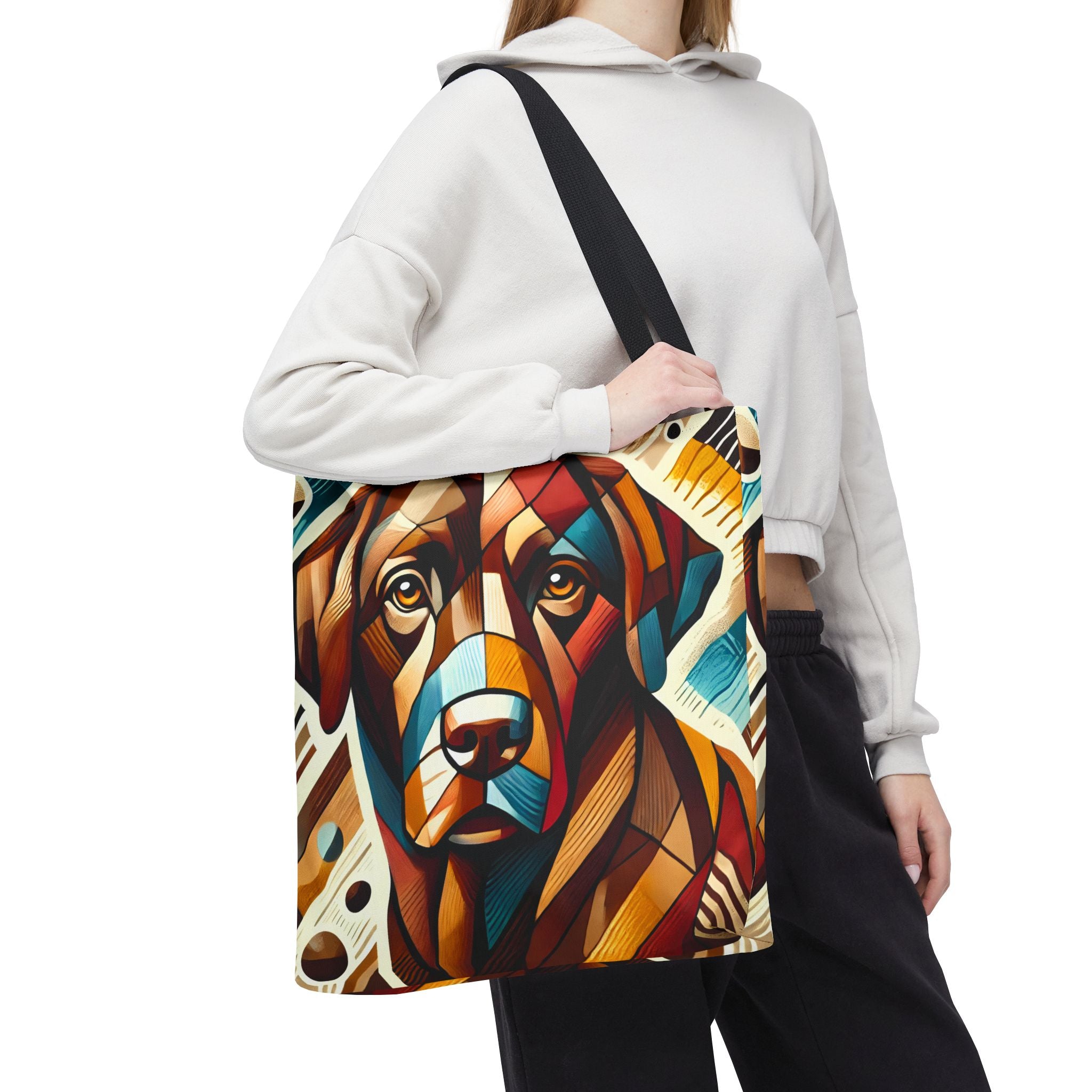 A person  carries a large tote bag with a colorful geometric chocolate Labrador retriever portrait print. The bag has a black shoulder strap and is shown against a plain white background.