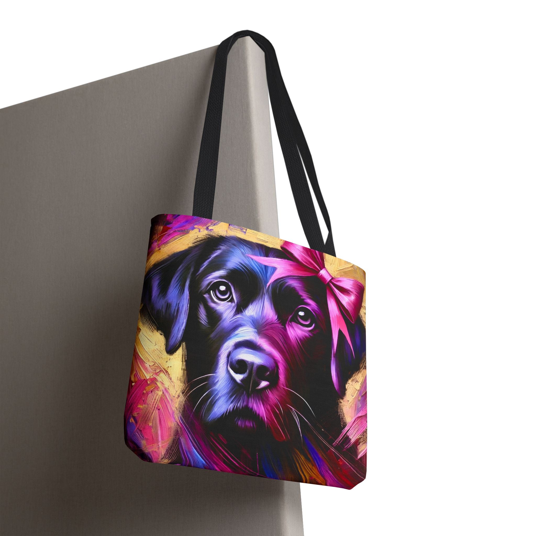 A tote bag with black shoulder straps, featuring a colorful, stylized portrait of a black Labrador retriever wearing a pink bow, set against a gray display stand on a white background.