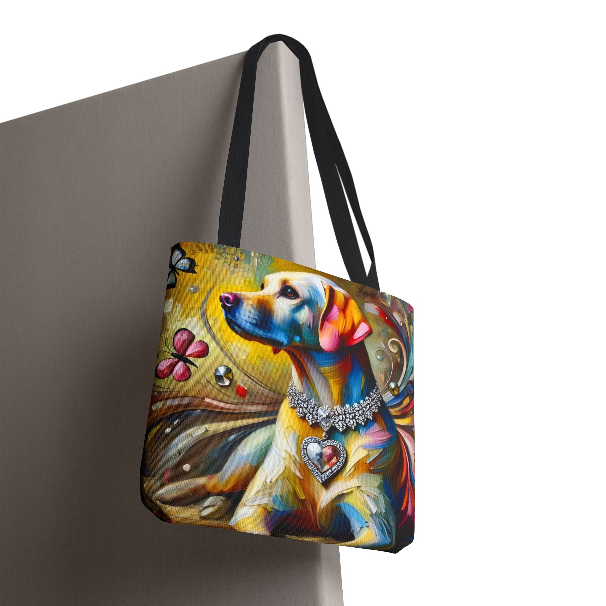 A tote bag with black shoulder straps, featuring a colorful artistic print of a yellow Labrador retriever wearing a heart-shaped collar, shown hanging against a neutral gray background.