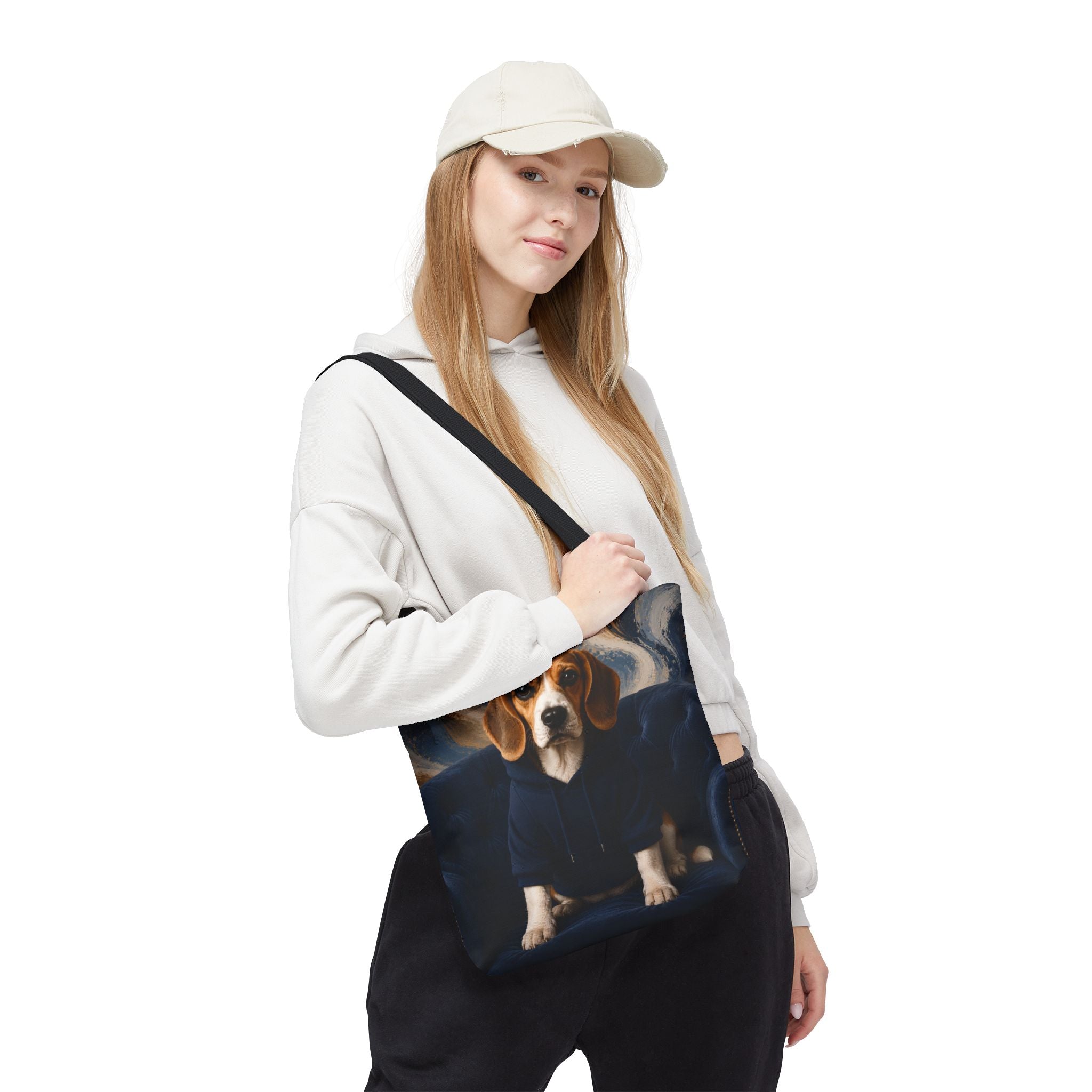 A woman wearing a beige cap, white hoodie, and black sweatpants carries a dark blue tote bag with a printed image of a beagle dog wearing a navy sweater. The background is solid white.