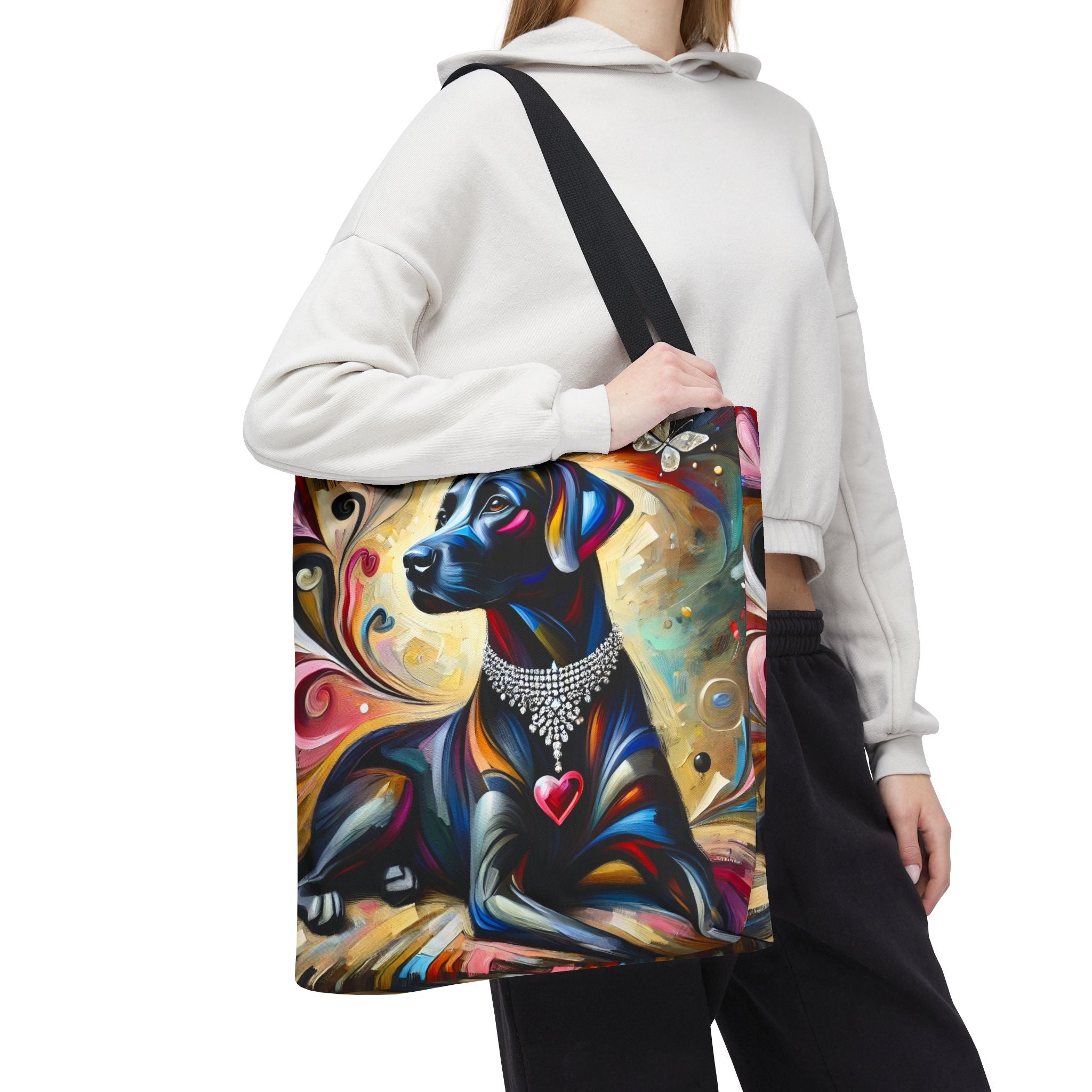 A person wearing a white cropped hoodie and black sweatpants carries a large tote bag with a colorful artistic print of a black Labrador retriever wearing a silver collar and heart pendant, against a plain white background.