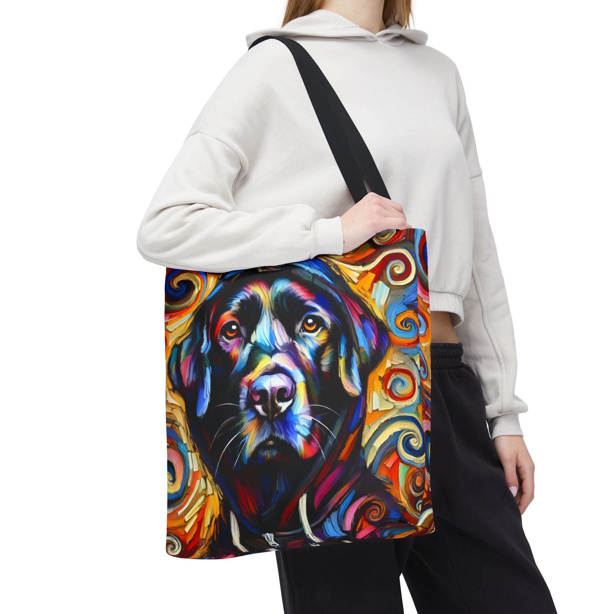 A person carries a large tote bag with a colorful painted black Labrador retriever portrait. The bag has a black shoulder strap and a vivid multicolor print featuring blue, yellow, orange, red, and black tones.
