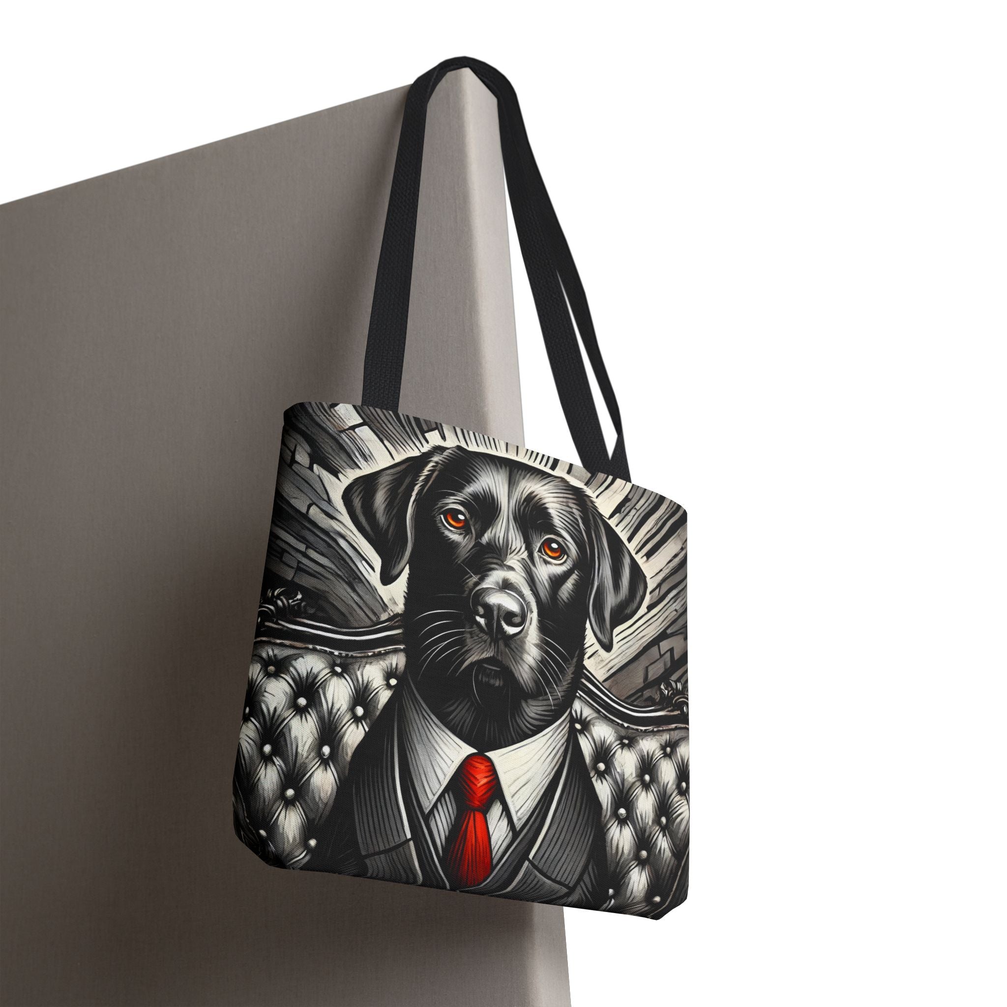 A tote bag with black handles, featuring a graphic print of a black Labrador Retriever dressed in a gray suit and white shirt with a bright red tie, hanging against a neutral gray background.