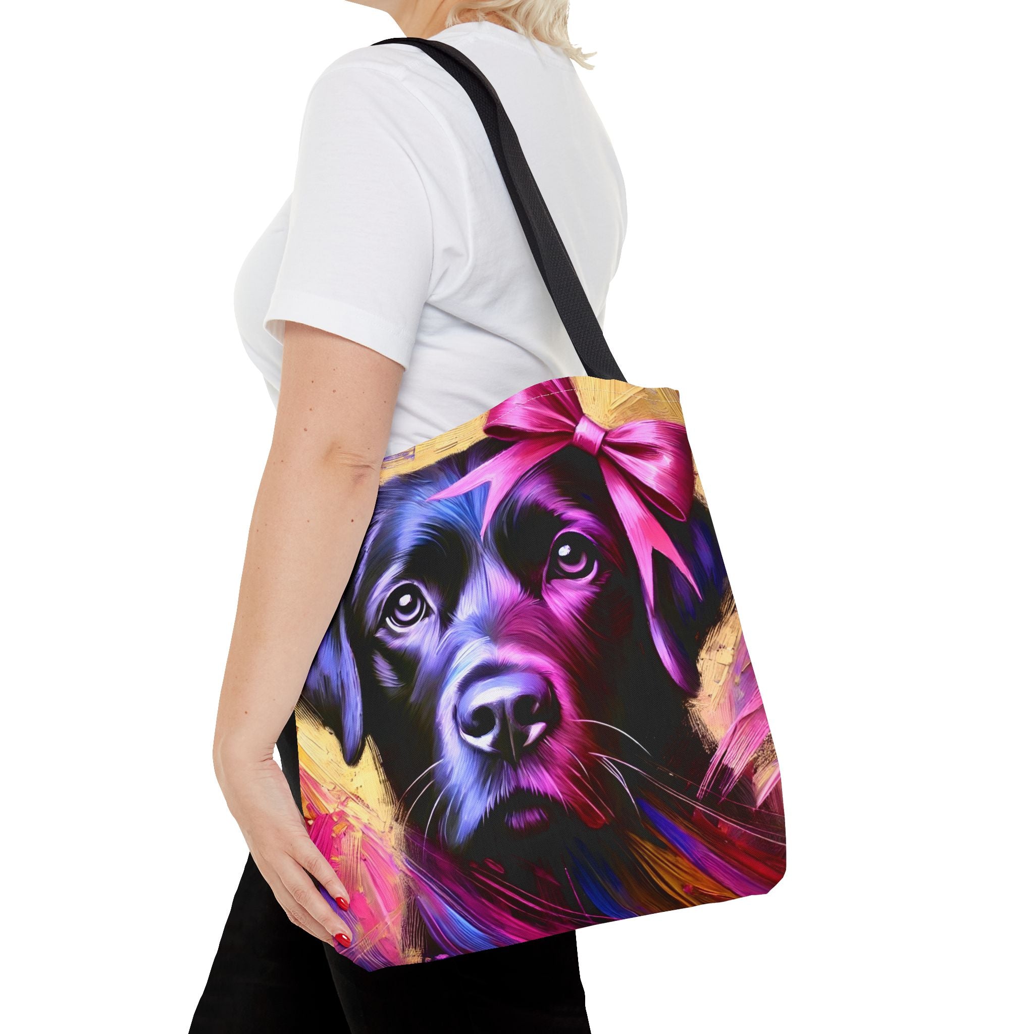 A woman carries a colorful tote bag featuring a stylized, vibrant portrait of a black Labrador with a pink bow. The bag has a black shoulder strap and is shown against a plain white background.