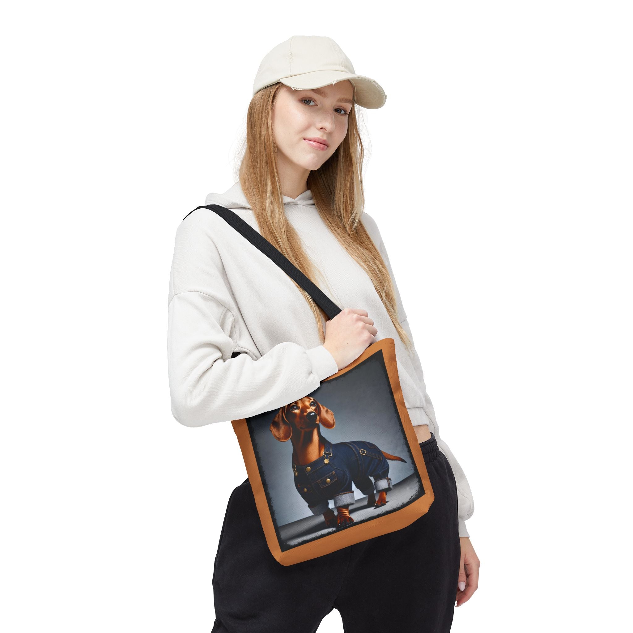 A woman wearing a beige cap, white hoodie, and black sweatpants carries a canvas tote bag with a brown leather-look trim and a printed graphic of a dachshund wearing blue jeans.
