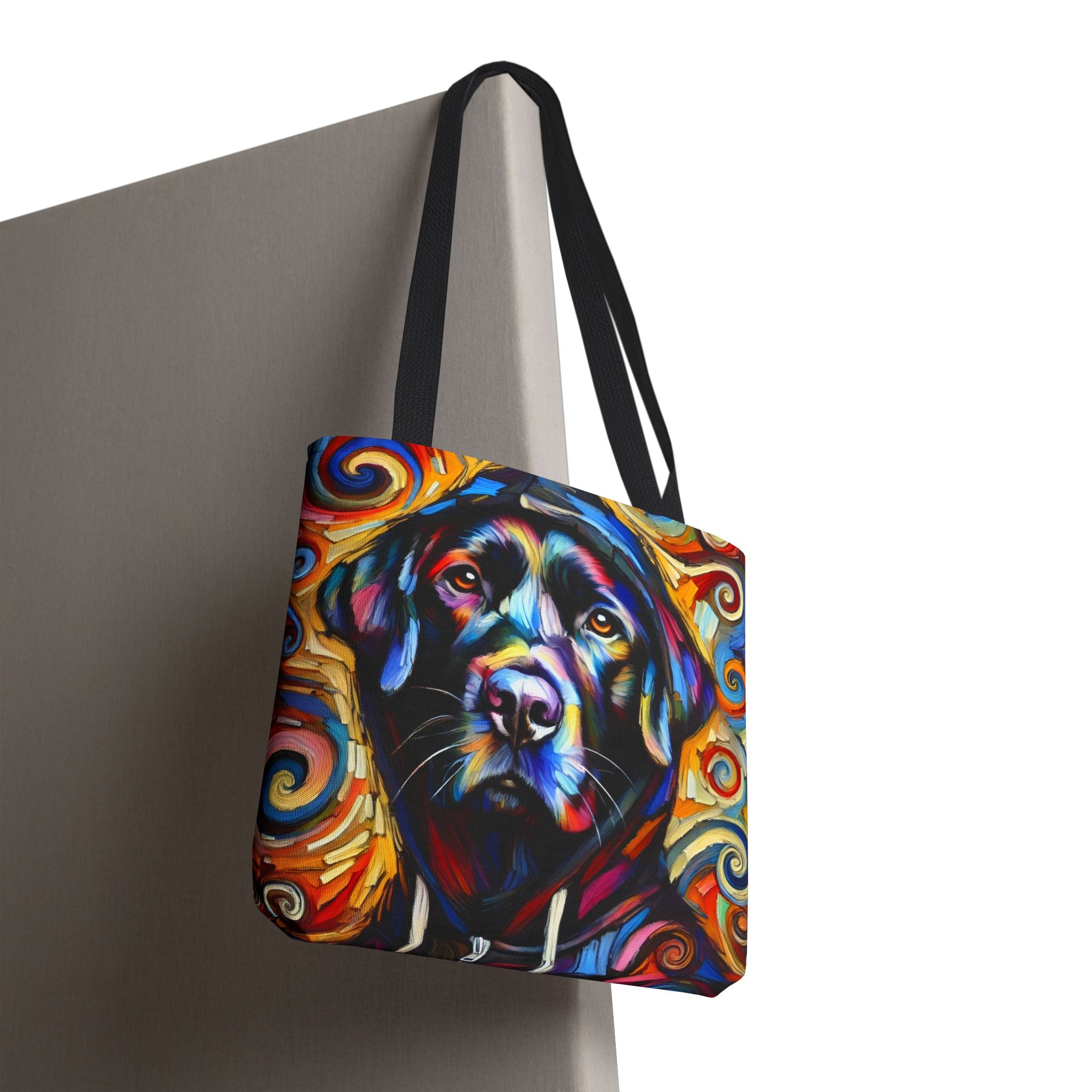 A tote bag with black shoulder straps, featuring a colorful, expressive painting of a black Labrador retriever surrounded by swirling abstract patterns in blue, yellow, orange, and red, shown hanging against a gray wall on a white background.