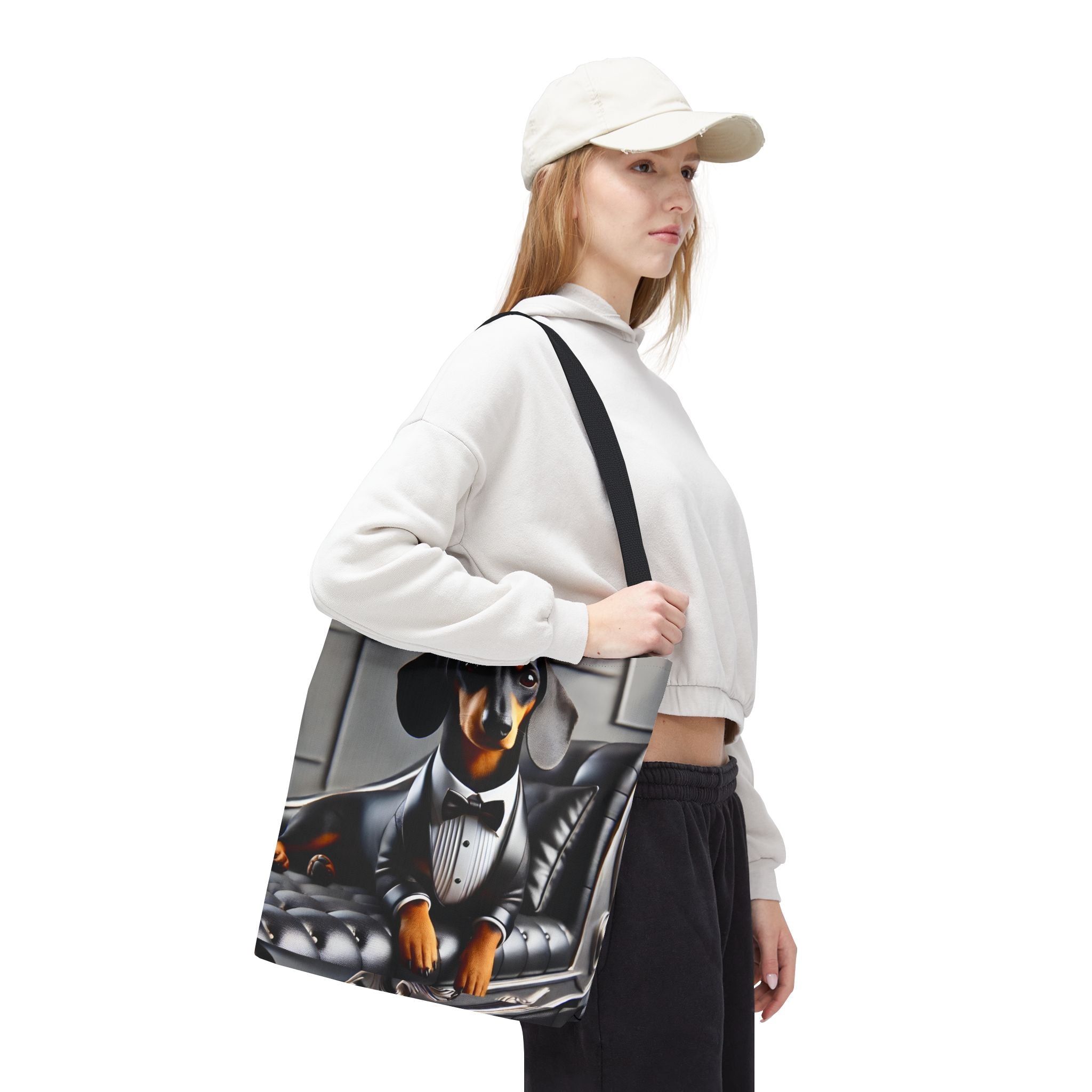 A woman wearing a white sweatshirt, black pants, and a white cap carries a canvas tote bag featuring a printed illustration of a dachshund in a tuxedo sitting in a car.
