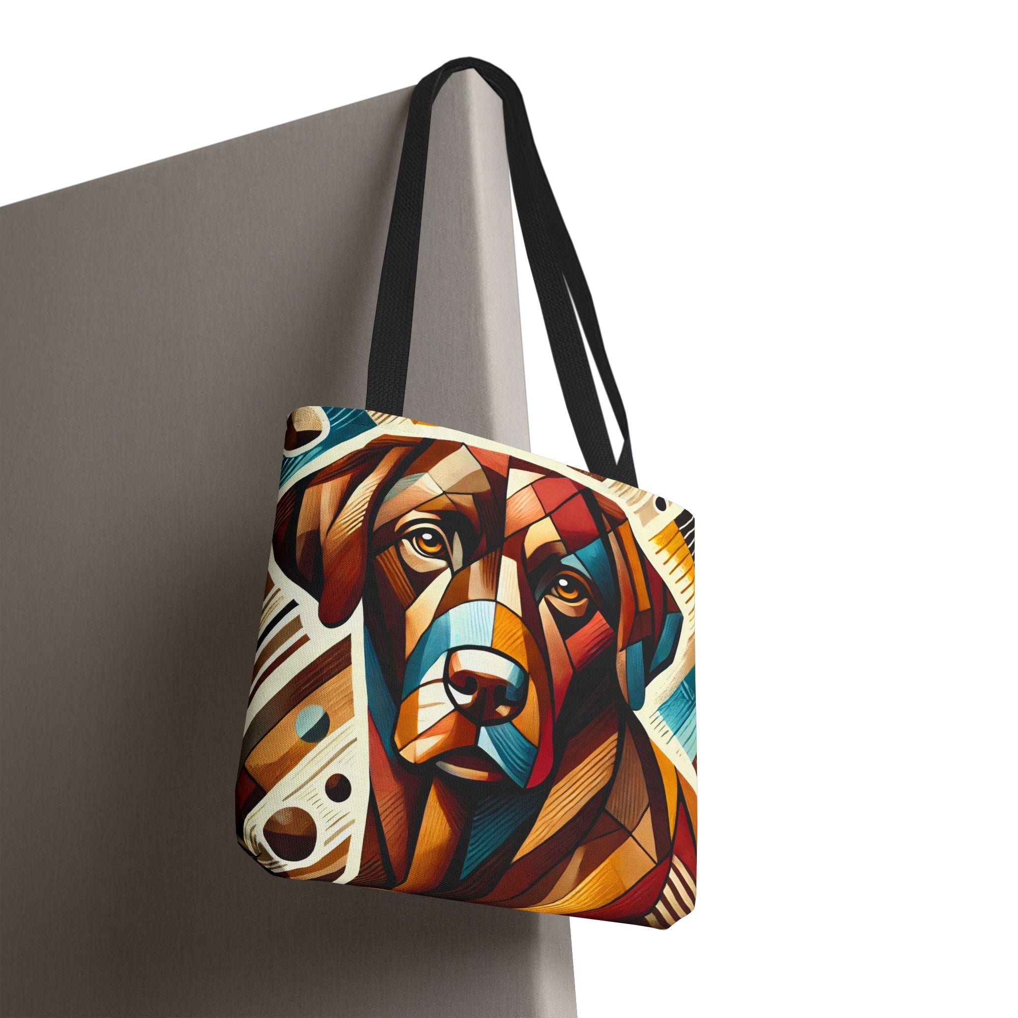 A tote bag with black shoulder straps, featuring a colorful geometric chocolate Labrador retriever portrait print, shown hanging against a gray wall on a white background.
