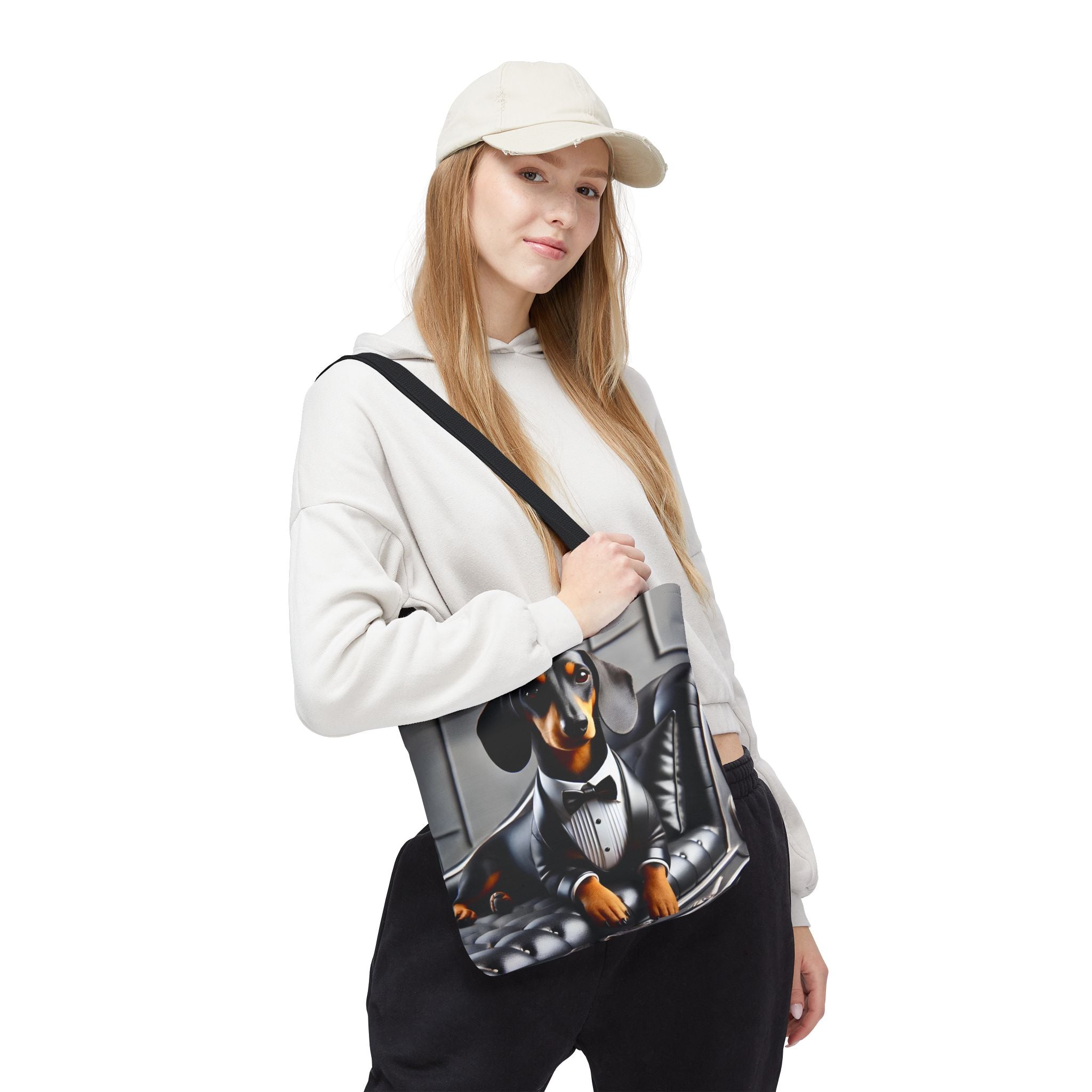 A woman wearing a beige cap, white hoodie, and black sweatpants carries a canvas tote bag featuring a printed graphic of a dachshund in a tuxedo. The background is solid white.